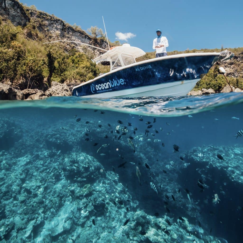 Fishing in Vanuatu September to December – Reef Edge and Centre Console | Ocean Blue Over‑under shot of an Ocean Blue centre‑console at a Vanuatu reef edge with schooling reef fish below, during the spring–summer fishing season.