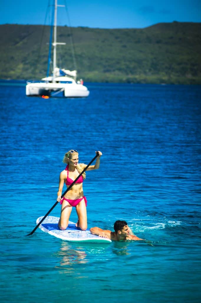 Couple enjoying stand‑up paddleboarding and a swim in clear blue water near a moored catamaran in Vanuatu.
