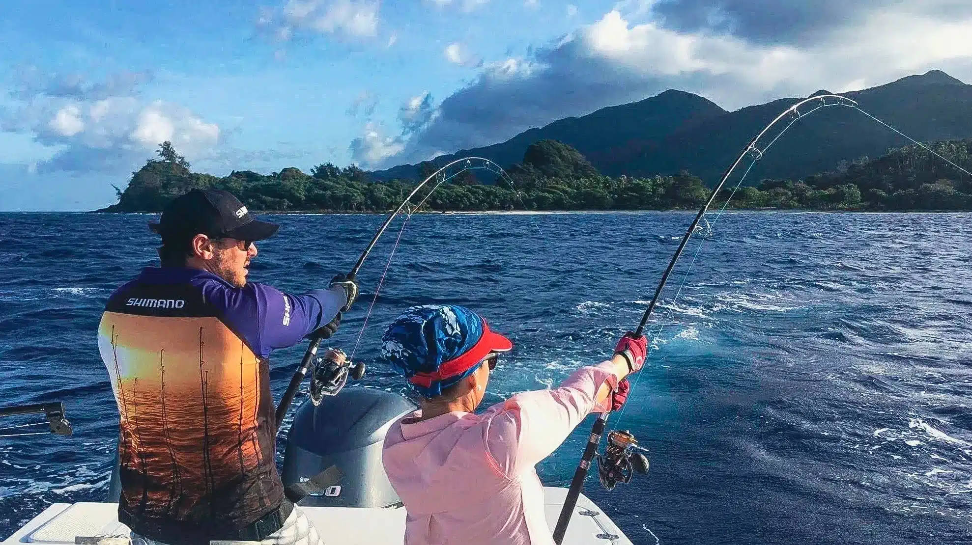 A couple sportfishing in Vanuatu on a boat, both holding bent fishing rods over deep blue water with tropical mountains behind.