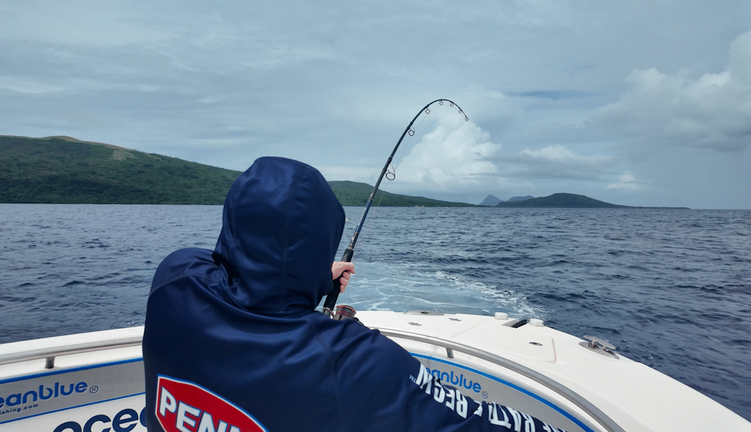 Angler fishing from an Ocean Blue Fishing Adventures boat in Vanuatu, rod bent with a fish on, open ocean and island coastline in the background.