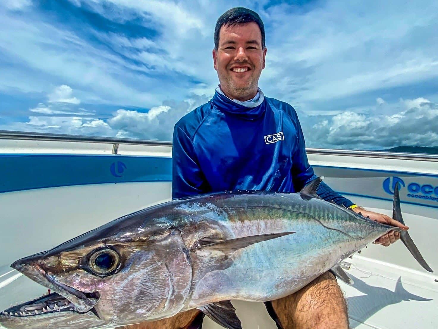 Smiling angler holding a large yellowfin tuna on an Ocean Blue Fishing Adventures boat in Vanuatu, with bright tropical sky and ocean in the background.