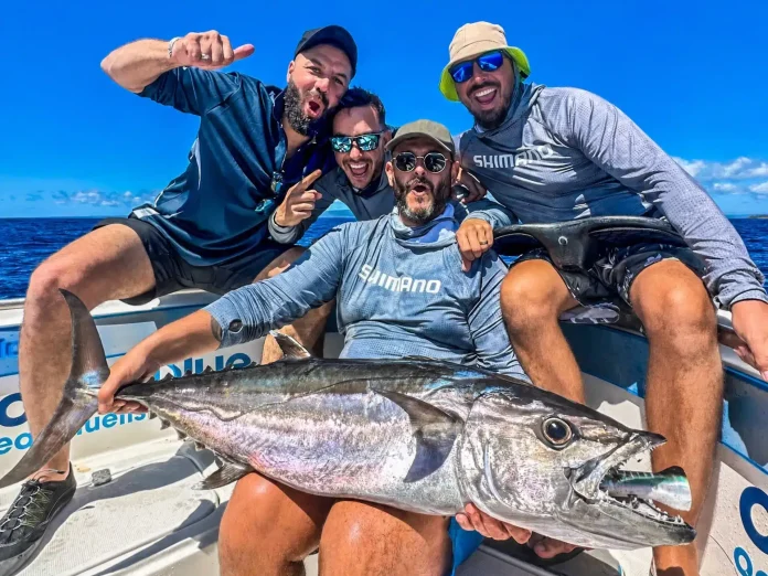 Four excited anglers on a fishing boat pose with a large dogtooth tuna, with open ocean and a clear blue sky in the background.