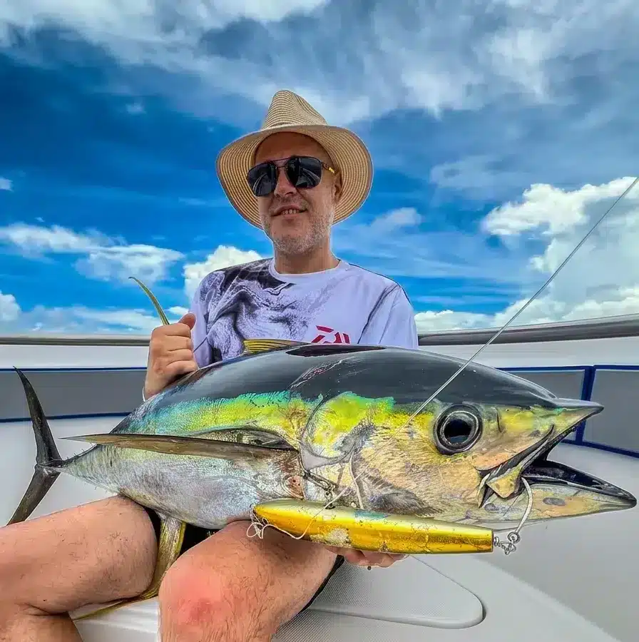Angler in a straw hat and sunglasses holds a large yellowfin tuna on a boat, with a lure and leader visible and a bright blue sky in the background.