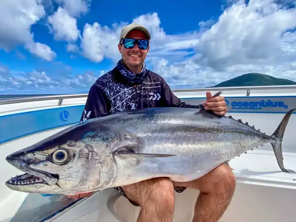 Smiling angler holding a large dogtooth tuna on an Ocean Blue Fishing Adventures boat in Vanuatu, with a bright blue sky, scattered clouds and island coastline in the background.
