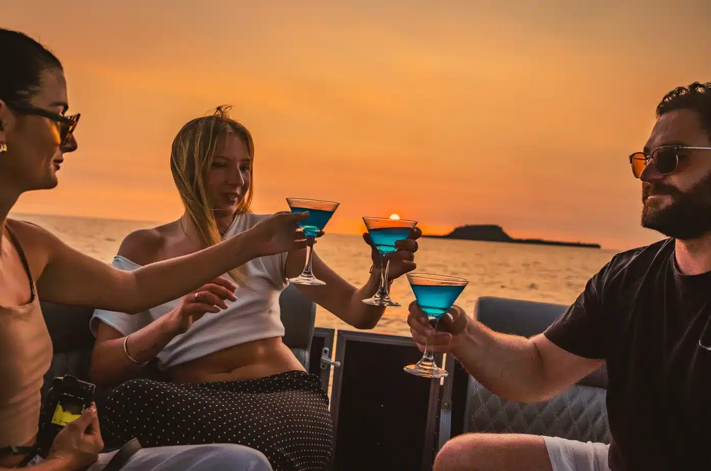 Guests clinking blue cocktails on a private sunset cruise near Trees and Fishes Private Retreat in Port Havannah, Vanuatu, with an orange sky, calm ocean and island silhouette at sunset.