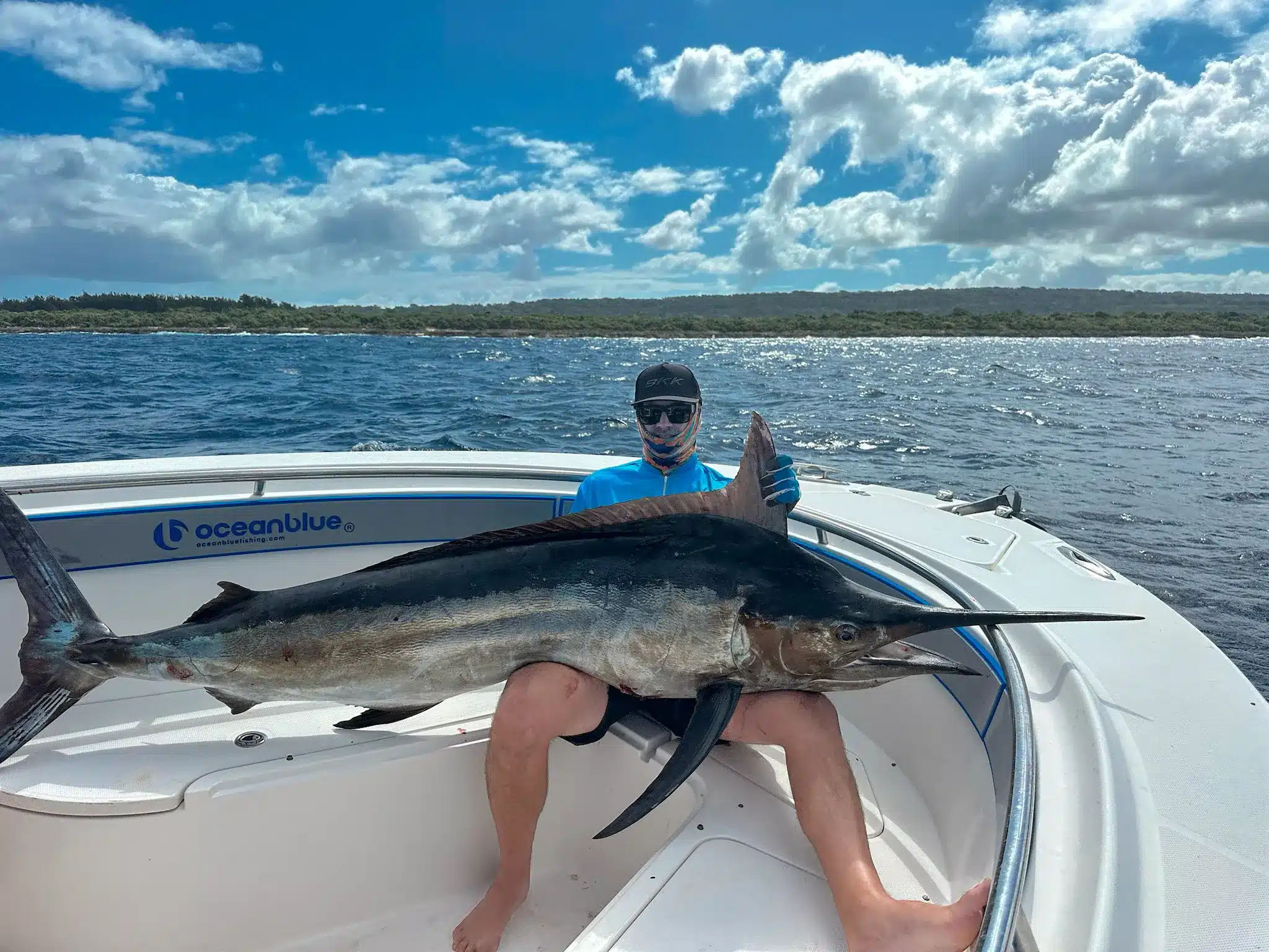 Angler holding a large swordfish on an Ocean Blue Fishing Adventures boat in Vanuatu, with choppy blue water and island coastline behind.