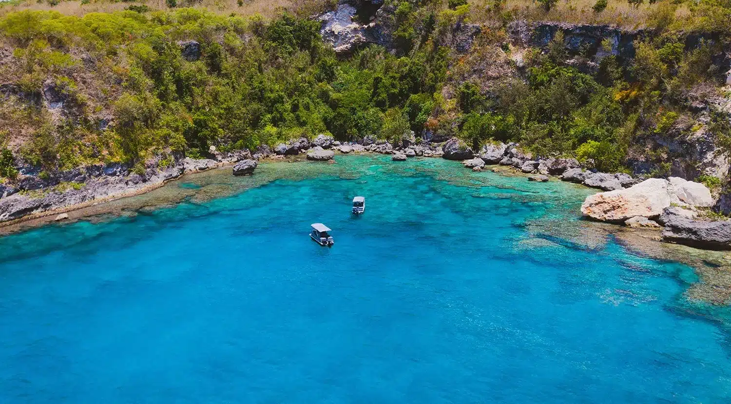 Aerial view of two boats anchored in Vanuatu's crystal-clear turquoise waters beside a rocky, tree-lined tropical coast