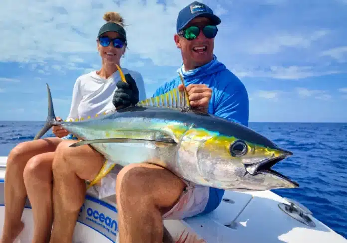 A couple sitting on a boat holding a large yellowfin tuna, smiling in bright sunshine on the ocean.