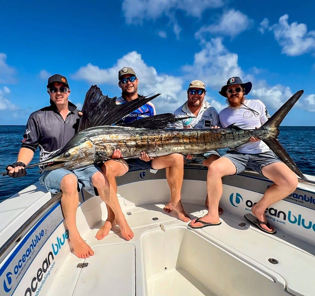 Four anglers holding a large sailfish on an Ocean Blue Fishing Adventures boat in Vanuatu, with blue sky and open ocean in the background.