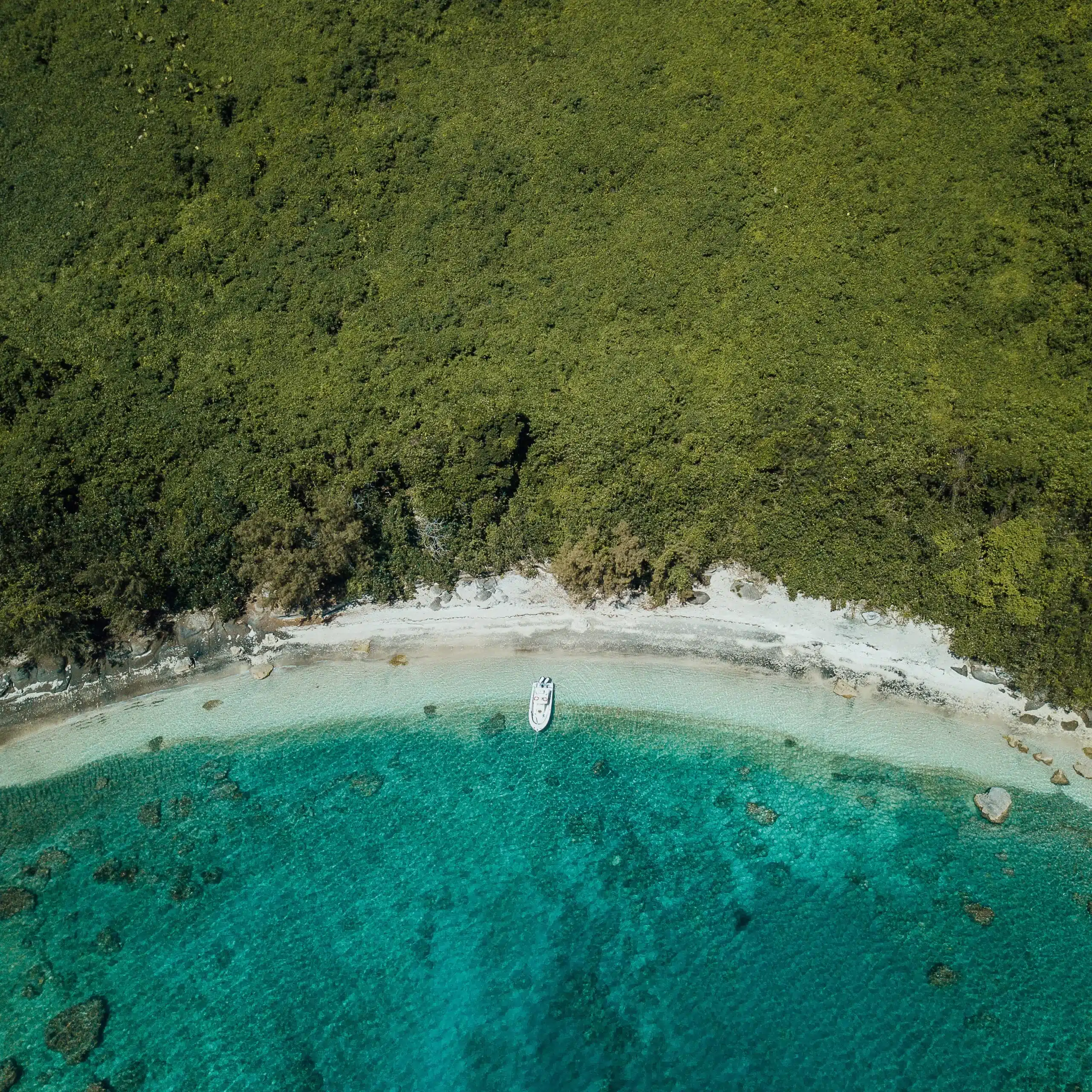 Drone view of a small boat anchored in clear turquoise water off a white sand beach with dense tropical rainforest behind in Vanuatu.