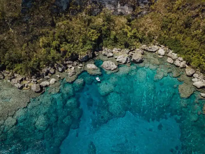 Aerial view of a small boat floating over a turquoise coral reef lagoon beside a rocky tropical shoreline in Vanuatu.