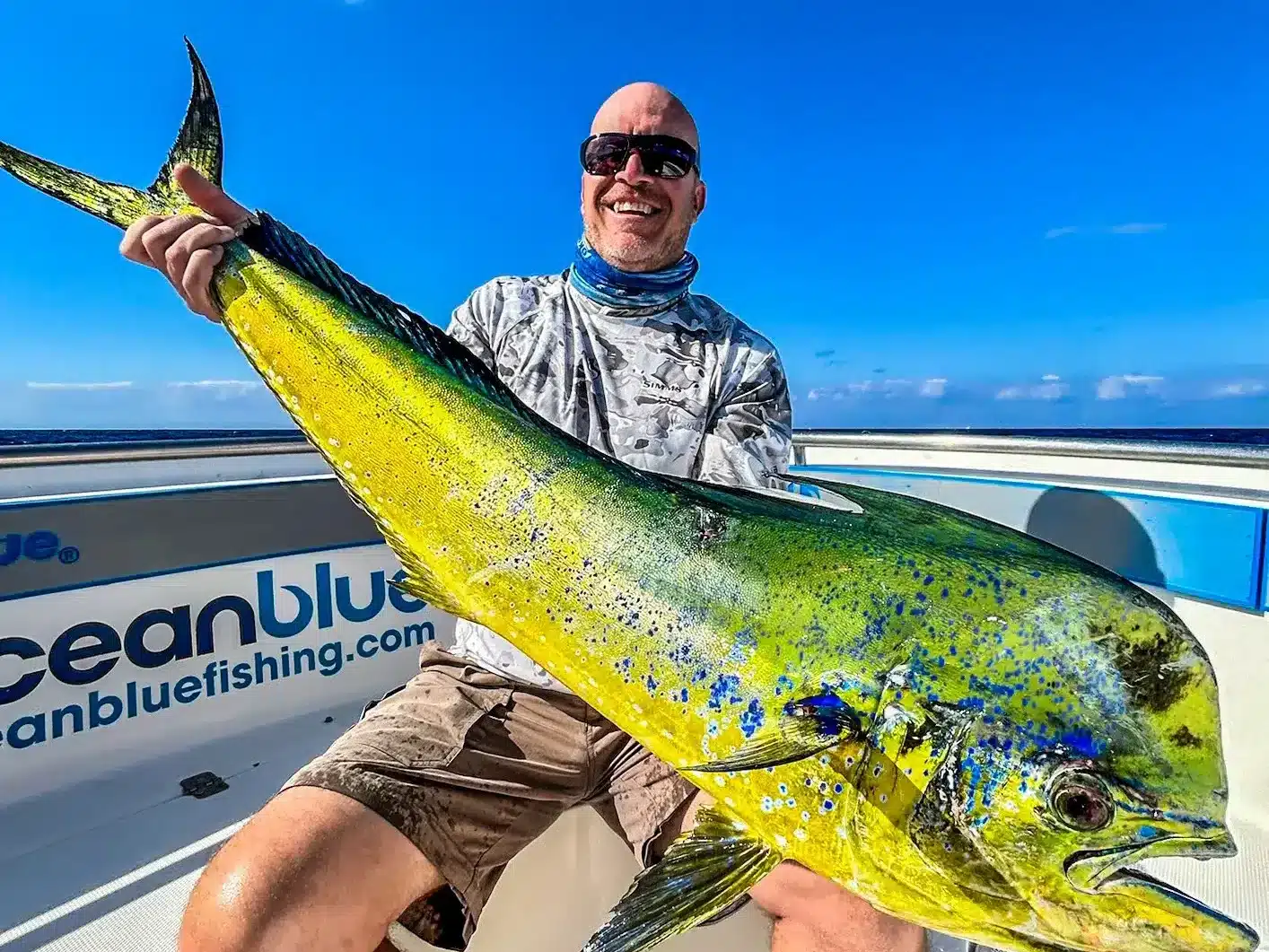 Smiling angler holding a large mahi mahi (dolphinfish) on an Ocean Blue Fishing Adventures boat in Vanuatu, with the open ocean and bright blue sky behind.