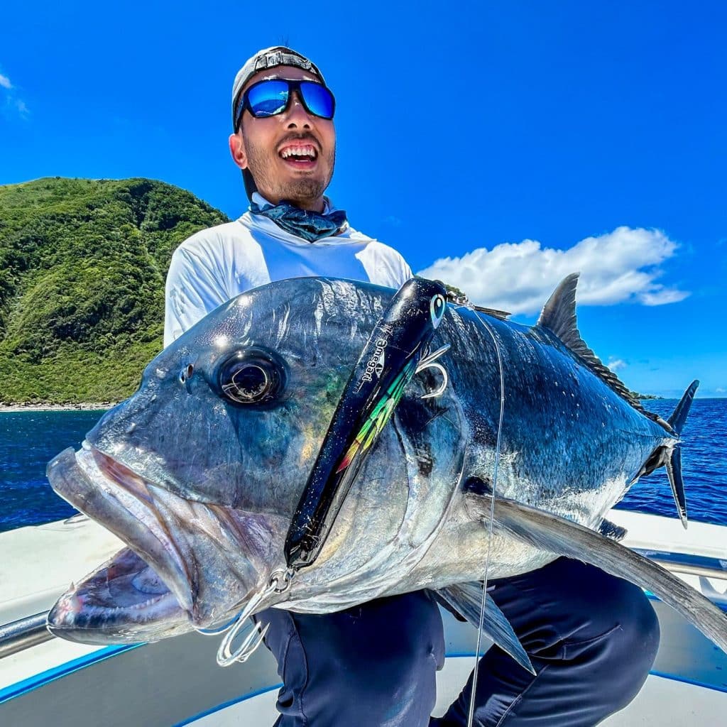 Angler holding a large giant trevally on a fishing boat in Vanuatu, with green island cliffs, deep blue ocean and a bright blue sky in the background.