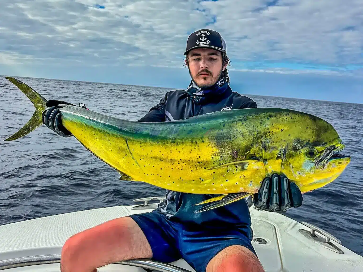 Angler holding a mahi mahi (dolphinfish) on a fishing boat in Vanuatu, with open ocean and cloudy sky in the background.