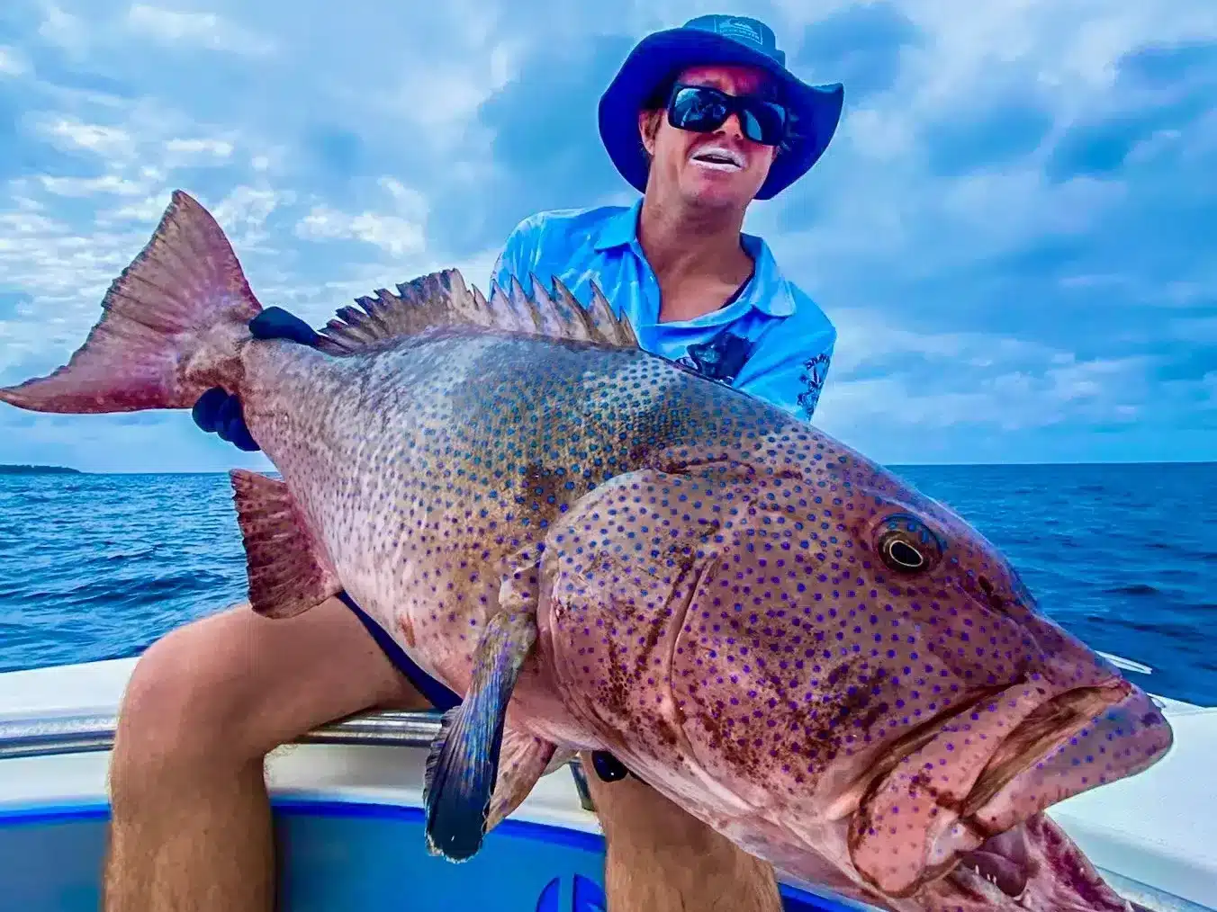 Angler wearing a wide-brim hat holding a large coral trout (coral grouper) on a fishing boat in Vanuatu, with calm ocean and cloudy sky in the background.