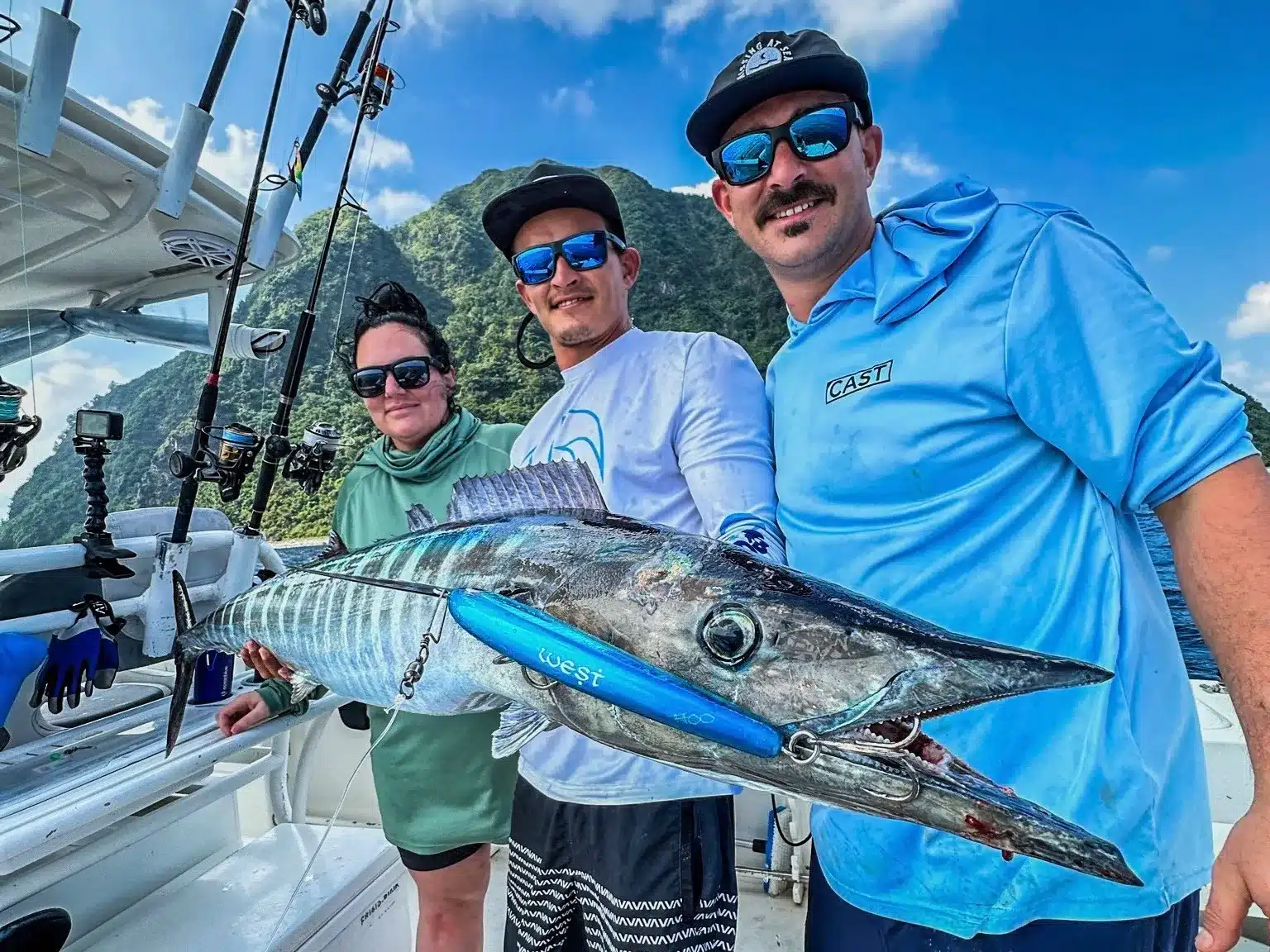 Three anglers posing with a large wahoo on a sportfishing boat in Vanuatu, with fishing rods, a bright blue lure and steep green island cliffs in the background.