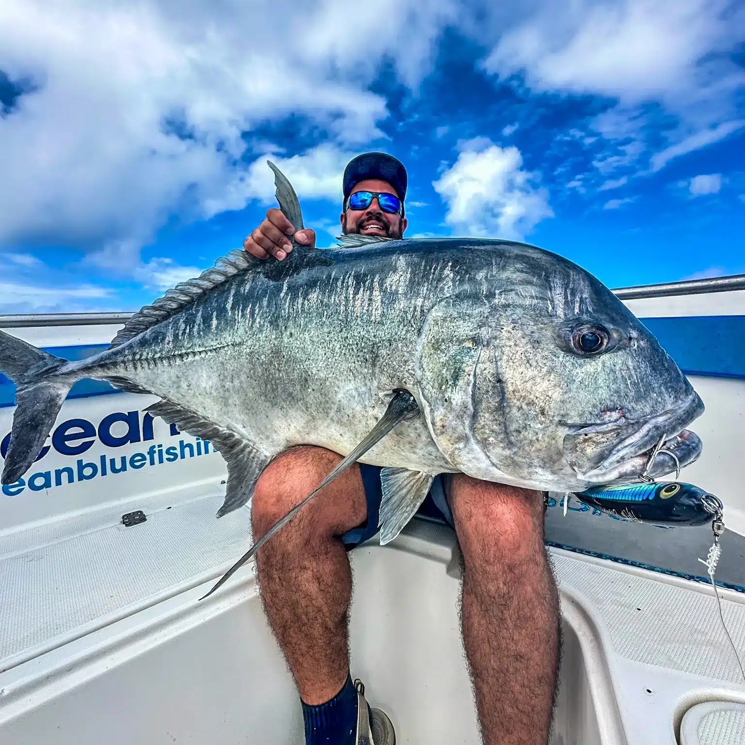 Angler holding a giant trevally (GT) on an Ocean Blue Fishing Adventures boat in Vanuatu, with blue sky and clouds overhead.