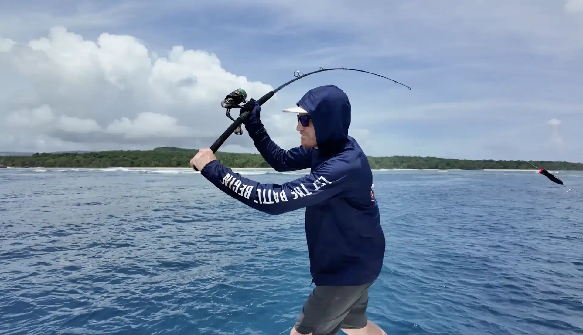 Angler casting a spinning rod from a boat on the ocean in Vanuatu, wearing a hooded fishing shirt with island coastline in the background.