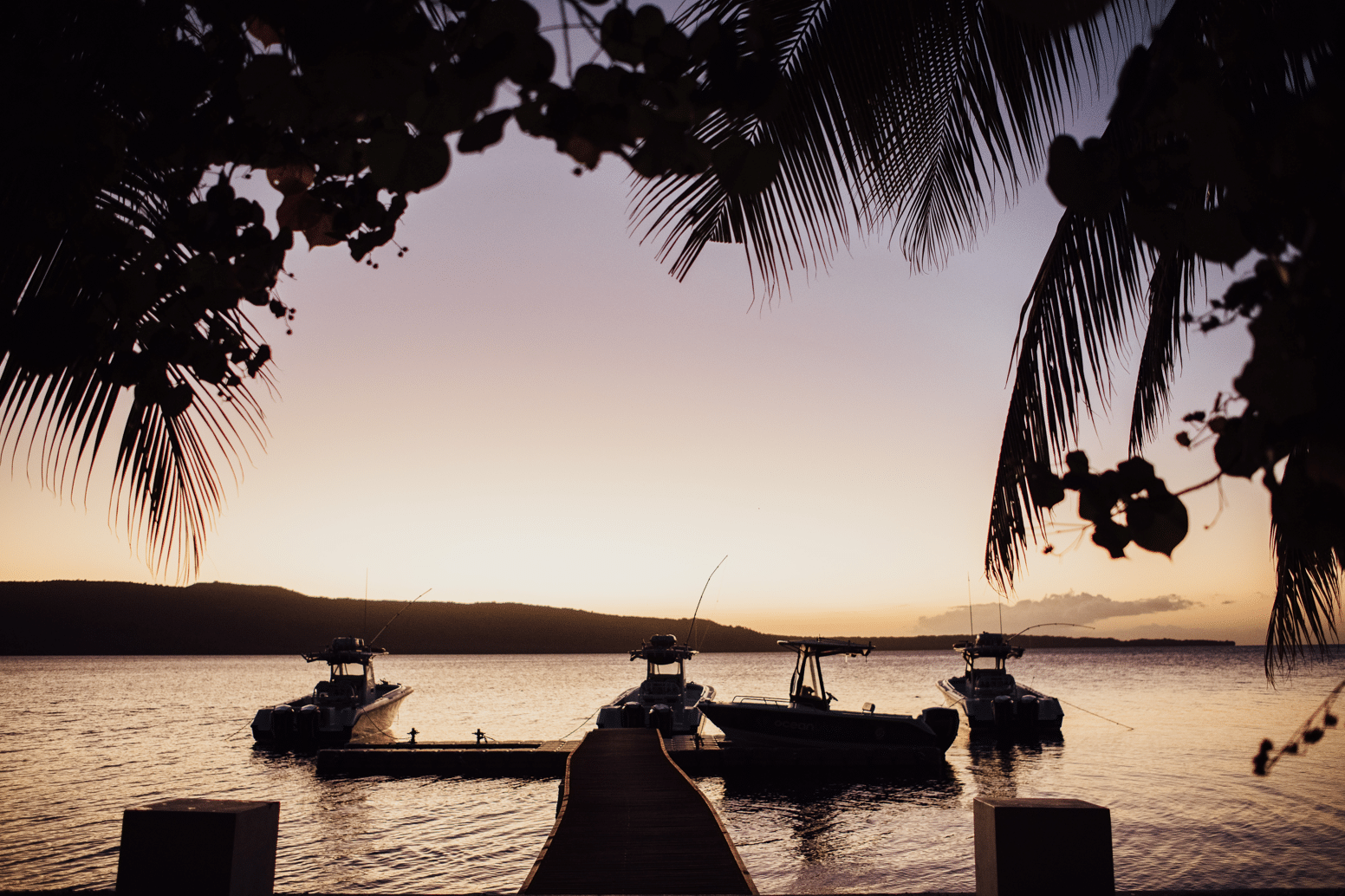Silhouette of sport fishing boats moored at a jetty in Vanuatu at sunset, framed by palm trees and calm ocean water.
