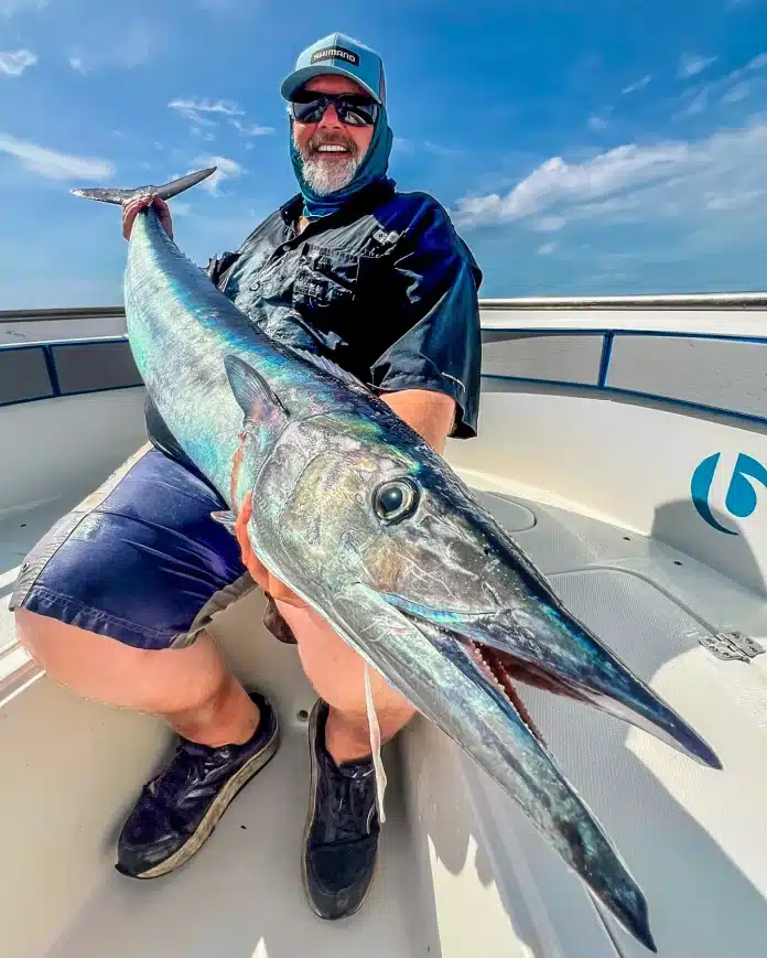 Angler holding a large wahoo on an Ocean Blue Fishing Adventures boat in Vanuatu, with blue sky and ocean in the background.