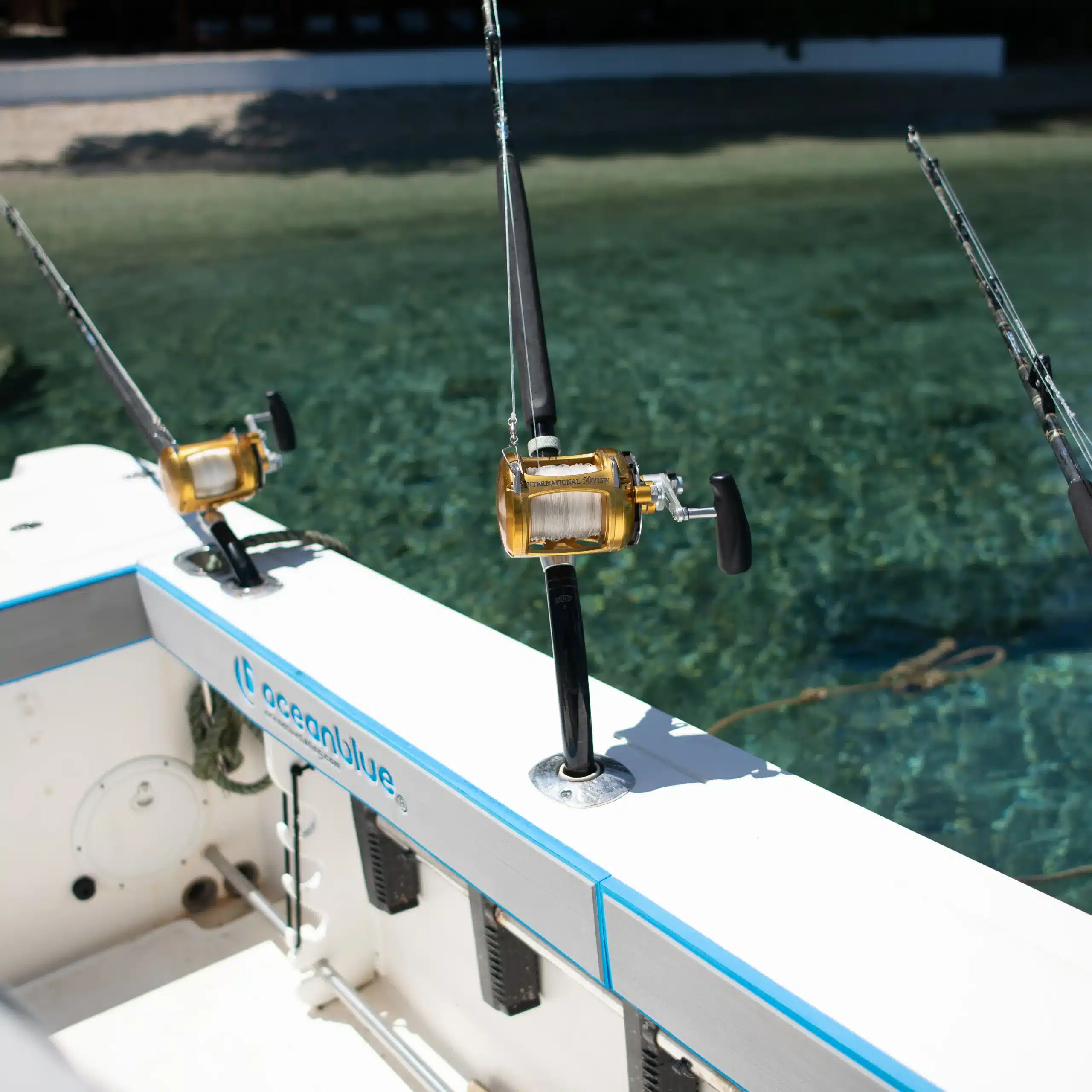 Close-up of trolling rods with gold game reels rigged on an Ocean Blue Fishing Adventures boat, with clear tropical water in the background.