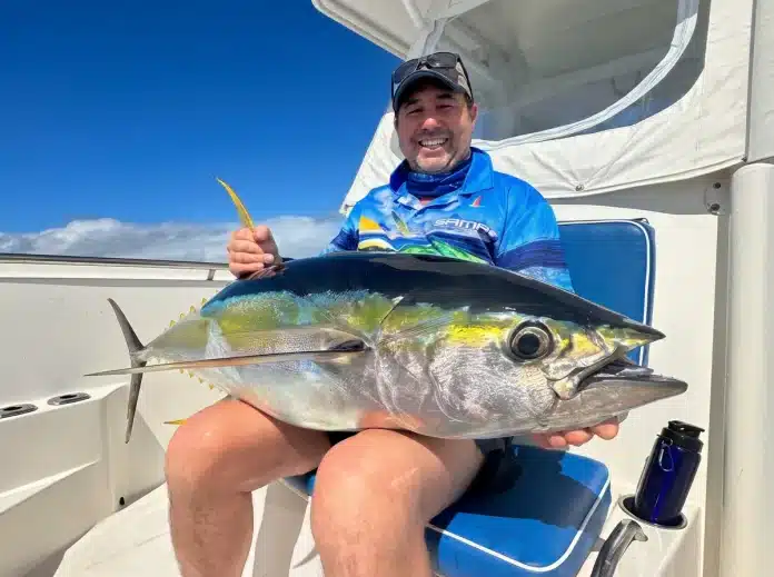 Smiling angler holding a yellowfin tuna on a sport fishing boat in Vanuatu, seated on the deck with blue sky behind.