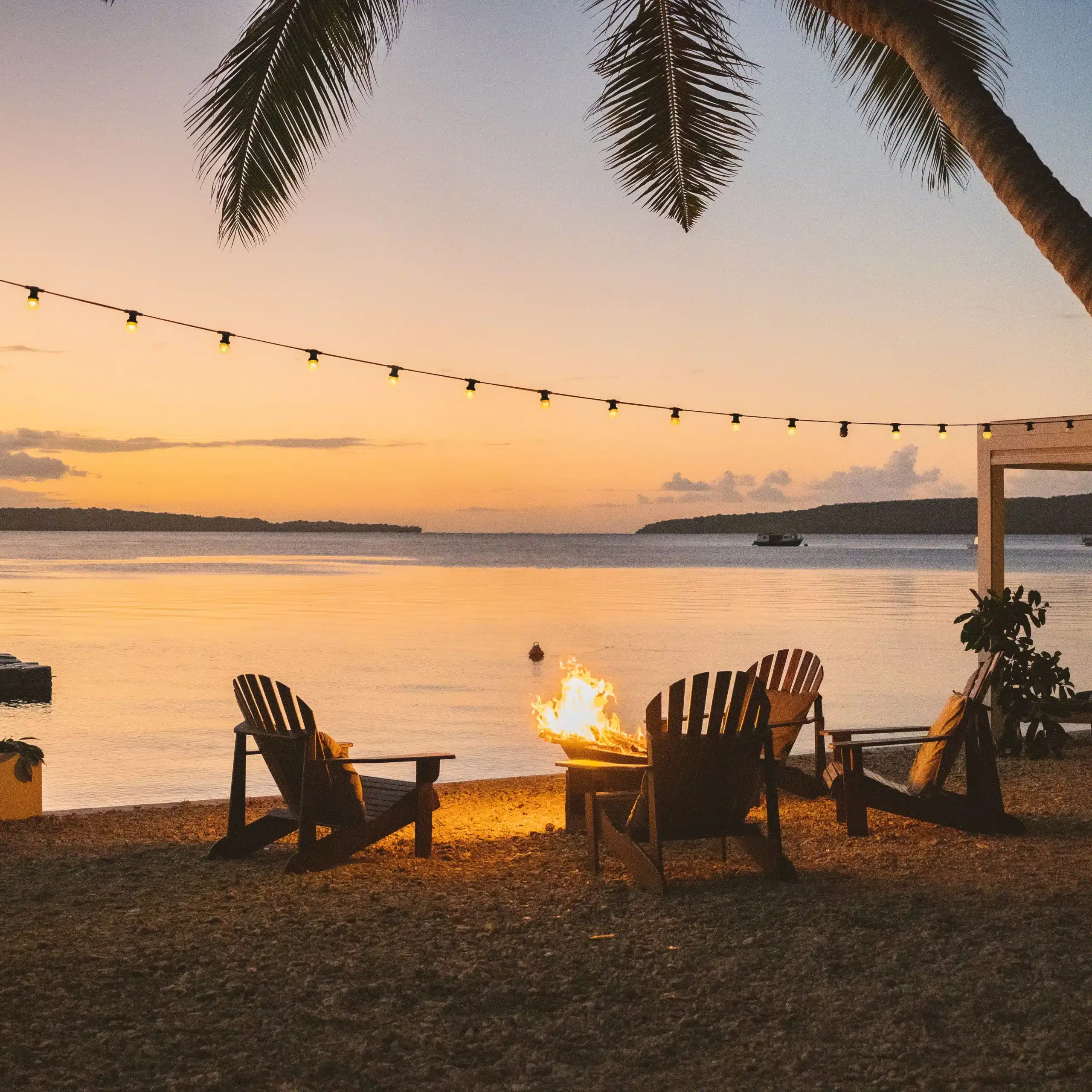 Beachfront fire pit with Adirondack chairs at sunset in Vanuatu, with string lights, calm water and palm trees in the background.