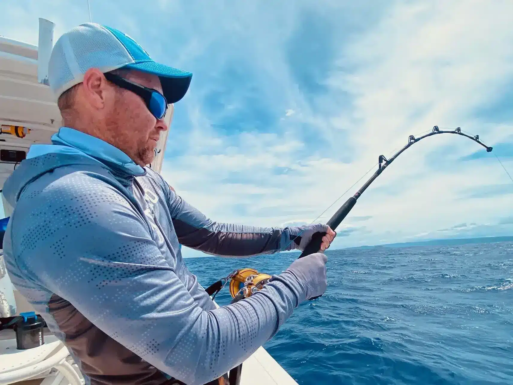 Angler fighting a fish with a bent rod and overhead reel on a sport fishing boat in Vanuatu, wearing sun-protective clothing on open ocean.