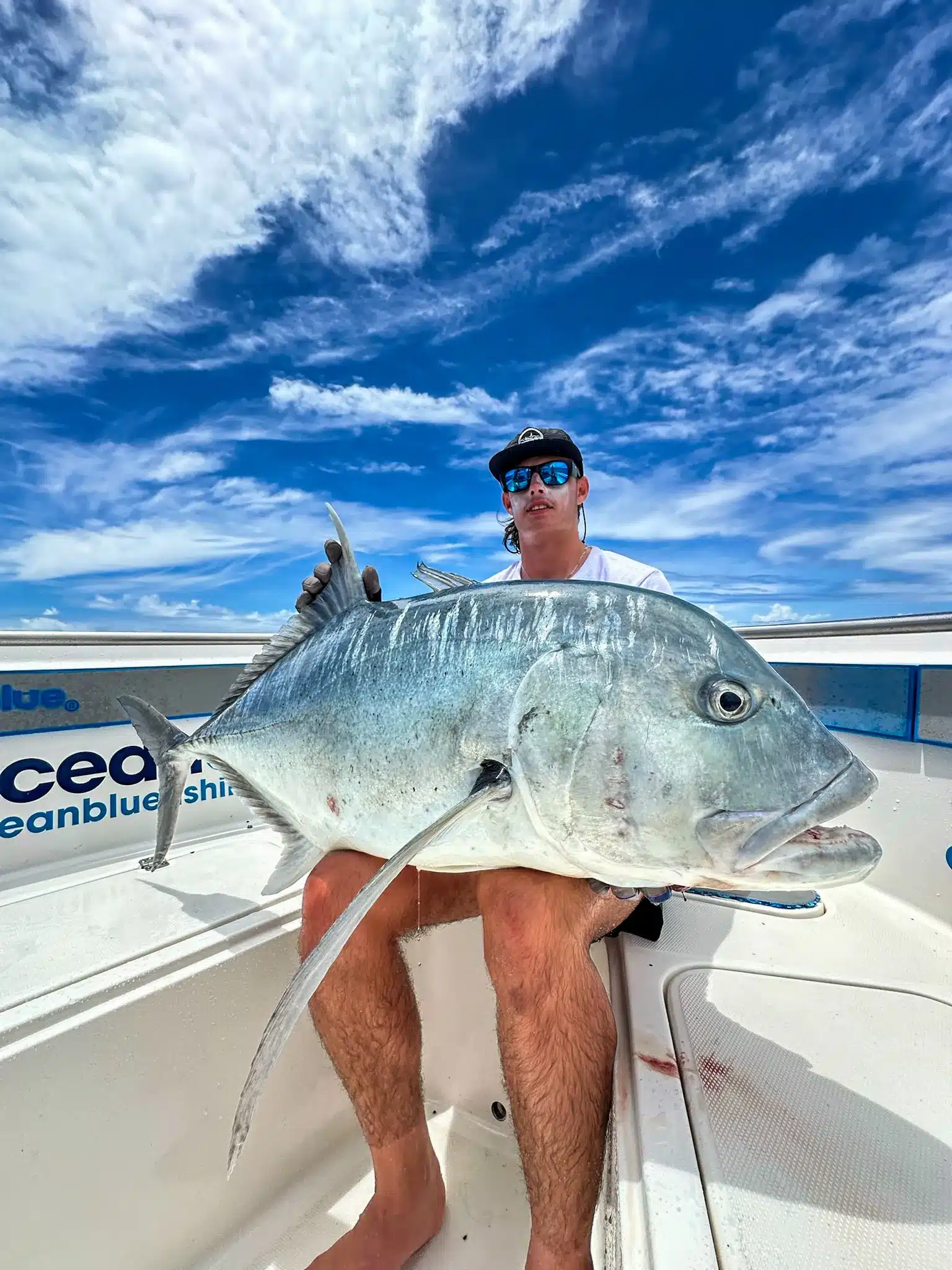 Angler wearing sun-protective fishing gear holding a large giant trevally (GT) on an Ocean Blue Fishing Adventures boat in Vanuatu under a bright blue sky.