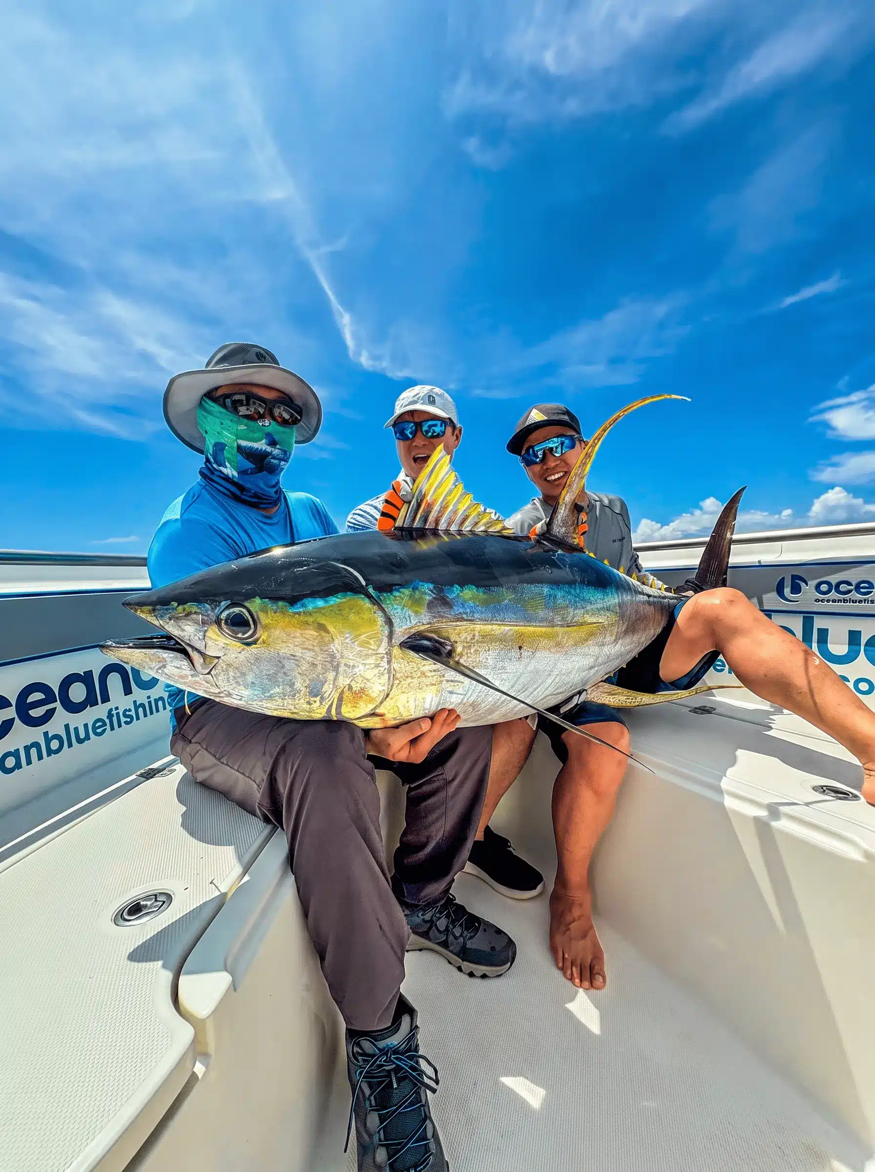 Three anglers wearing sun-protective fishing clothing holding a large yellowfin tuna on an Ocean Blue Fishing Adventures boat in Vanuatu under a bright blue sky.