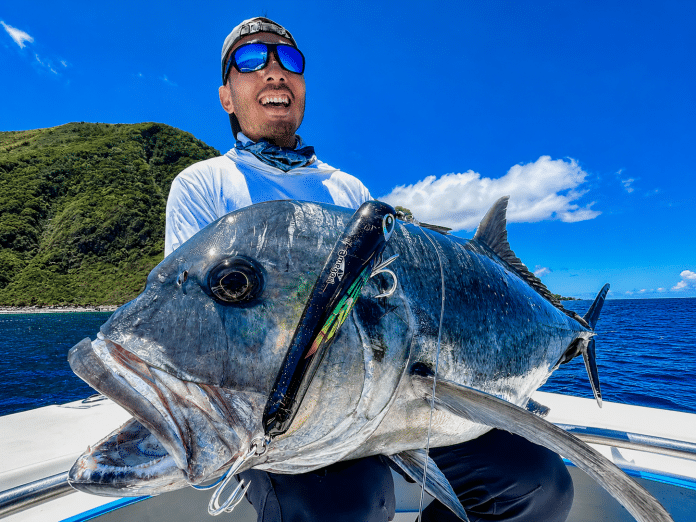 Angler holding a large giant trevally (GT) on a boat in Vanuatu, with a stickbait lure hooked in the fish’s mouth and a lush island coastline behind.