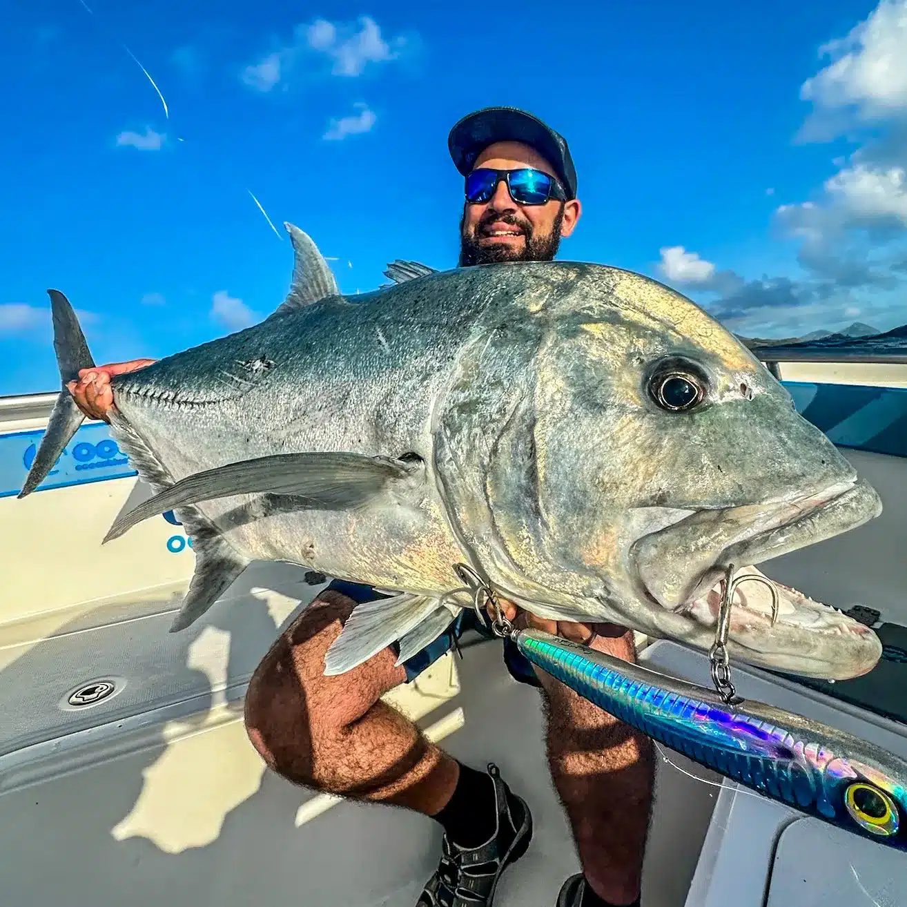Angler holding a large giant trevally (GT) on a boat in Vanuatu, with a metal jig lure in the fish’s mouth and blue sky behind.