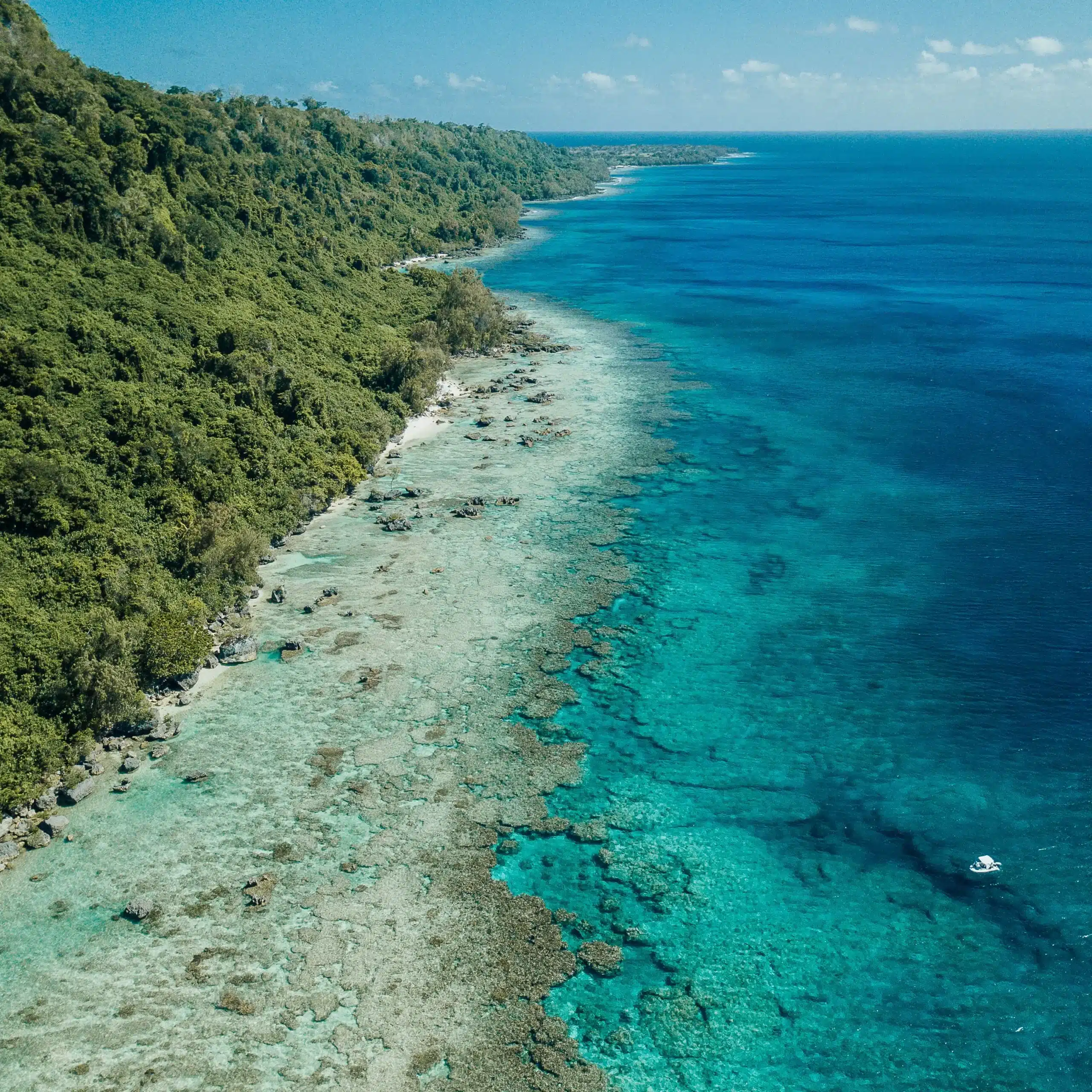 Aerial view of Vanuatu coastline with a shallow coral reef shelf and turquoise water fading into deep blue ocean, with a small boat offshore.