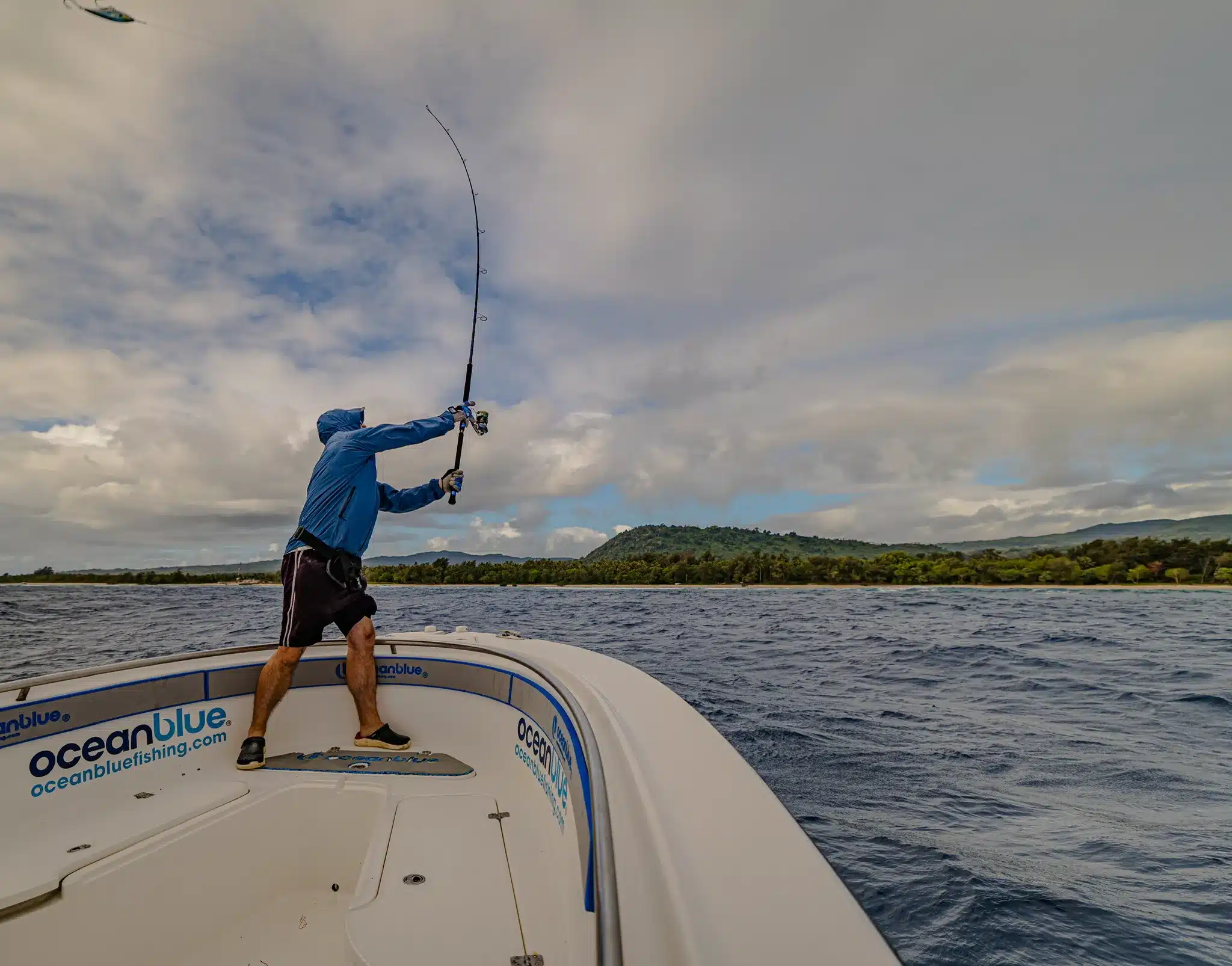 Angler casting a rod from the bow of an Ocean Blue Fishing Adventures boat in Vanuatu, with open water and island coastline under cloudy skies.