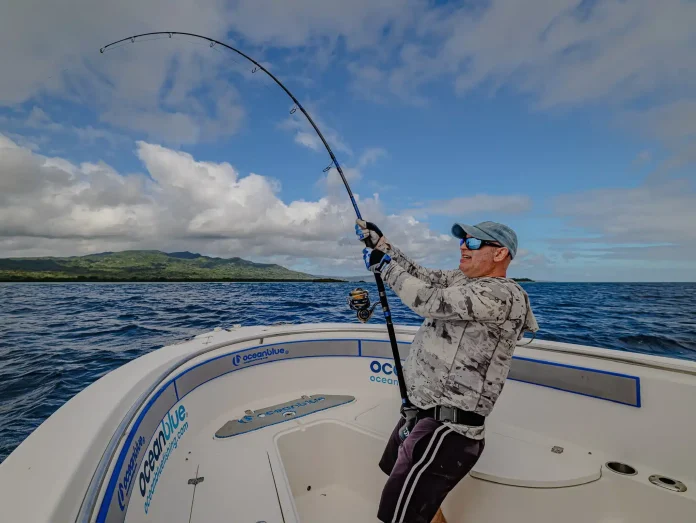 Angler fighting a strong fish with a bent rod on the bow of an Ocean Blue Fishing Adventures boat in Vanuatu, with island coastline in the background.