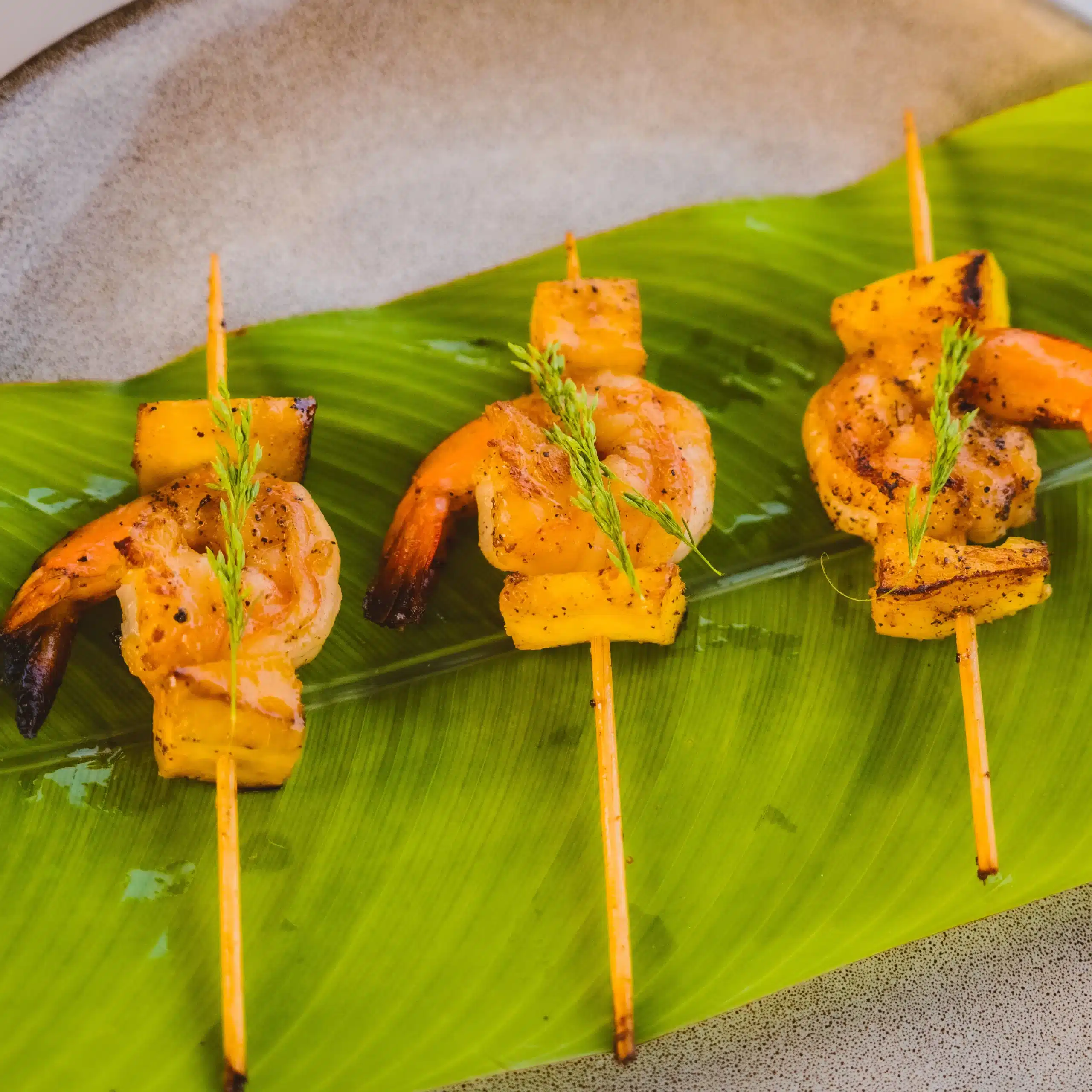 Grilled prawn skewers with pineapple pieces served on a banana leaf, garnished with herbs as gourmet canapés.