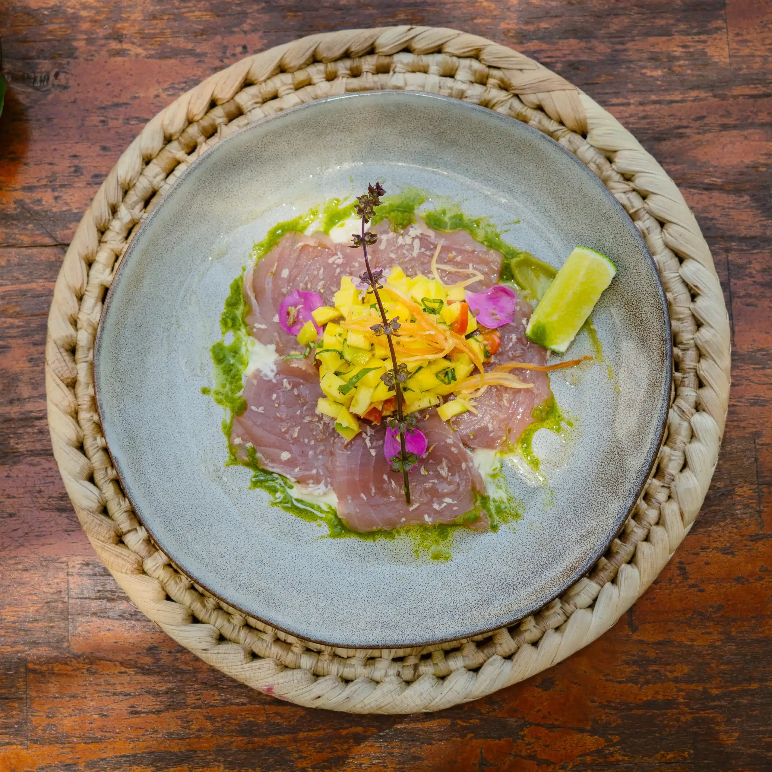 Top-down view of a plated raw fish dish with sliced fish, mango salsa, herbs and edible flowers, served with a lime wedge and green sauce.
