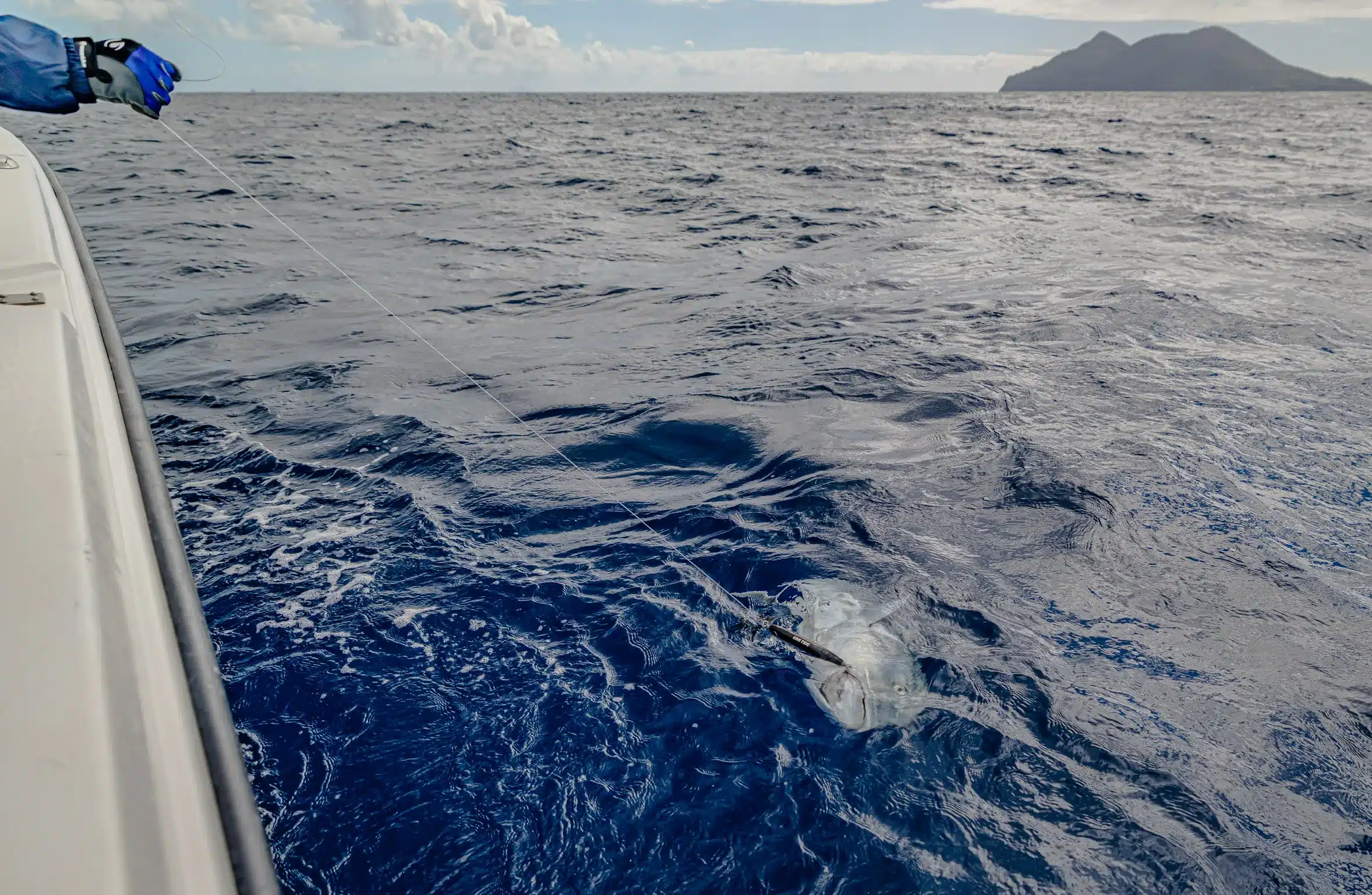 Angler’s gloved hand holding a leader line as a large fish is brought boatside in deep blue water off Vanuatu, with island coastline in the distance.