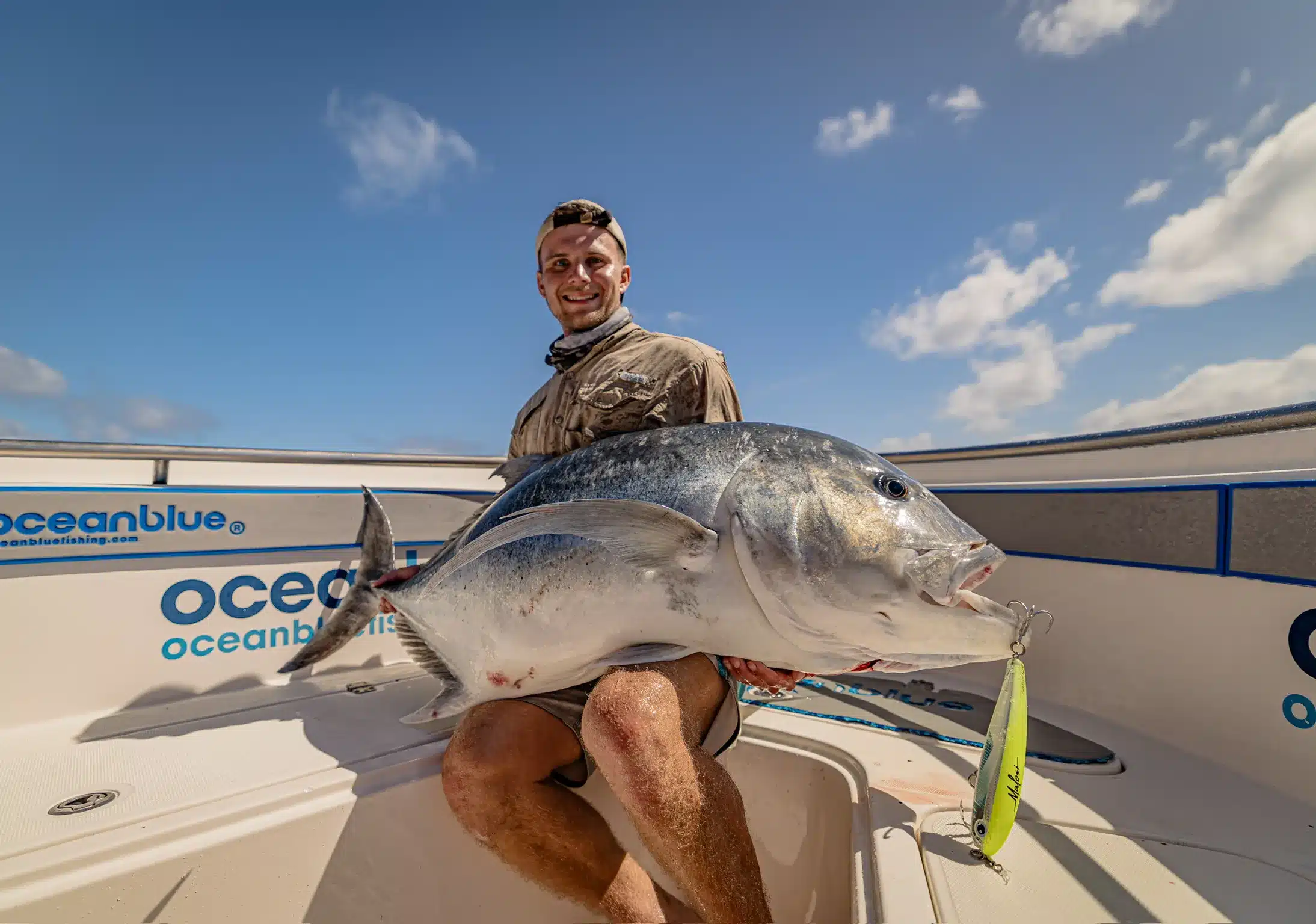 Angler holding a large giant trevally (GT) with a lure in its mouth on an Ocean Blue Fishing Adventures boat in Vanuatu, with the Ocean Blue logo visible.