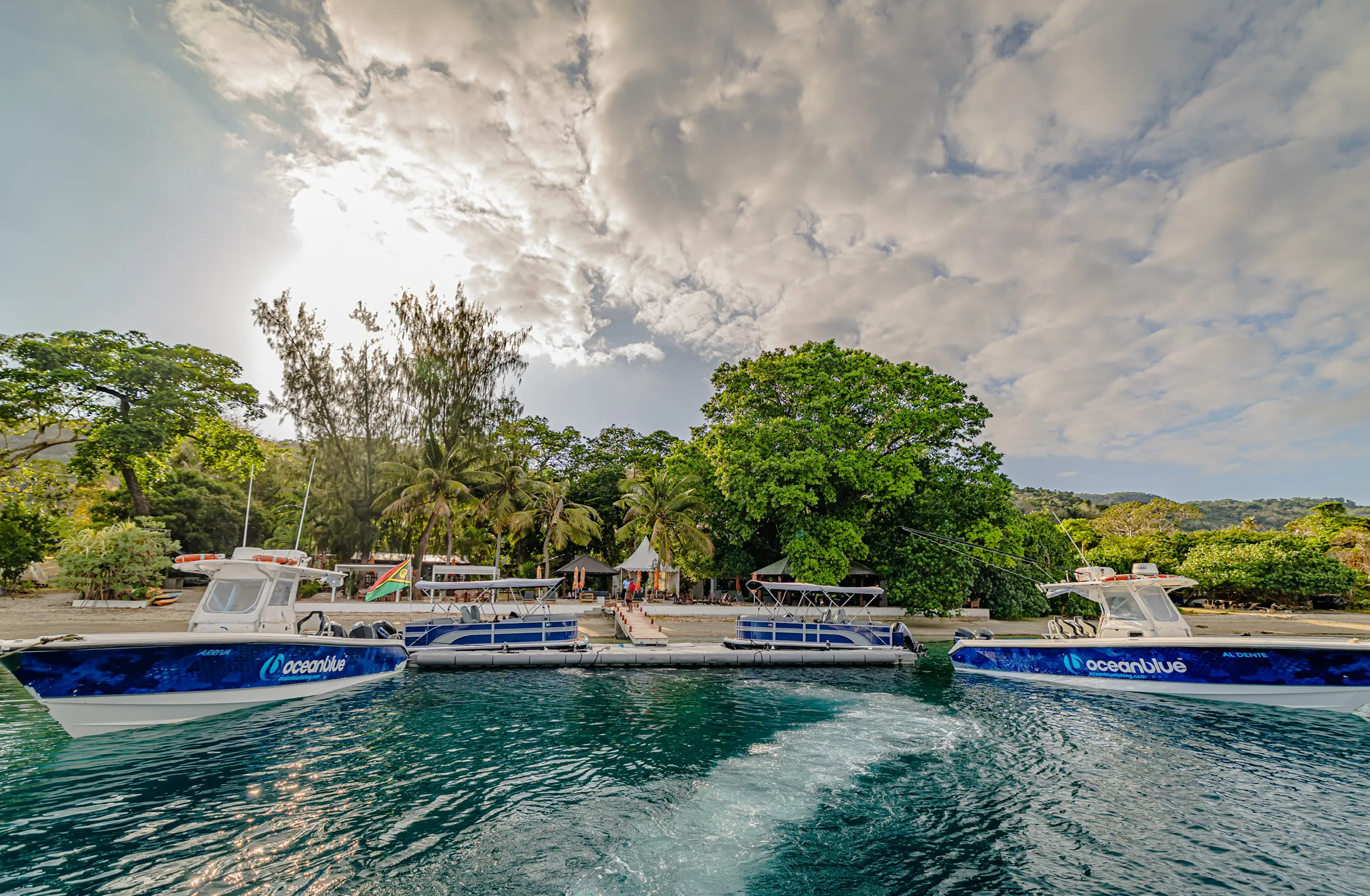 Ocean Blue Fishing Adventures boats moored at the jetty by the shoreline in Vanuatu, with tropical trees and the lodge area in the background.