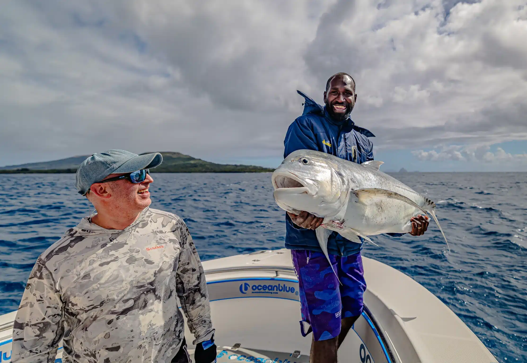 Ocean Blue Fishing Adventures crew member holding a large giant trevally (GT) on a boat in Vanuatu, with another angler on deck and island coastline behind.