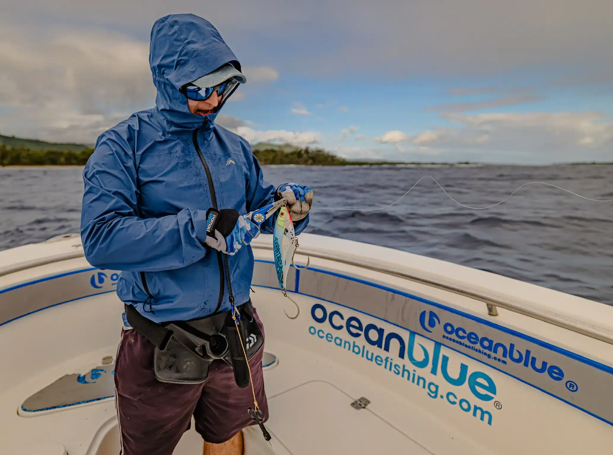 Angler in a rain jacket rigging a lure on an Ocean Blue Fishing Adventures boat in Vanuatu, with open ocean and coastline in the background.