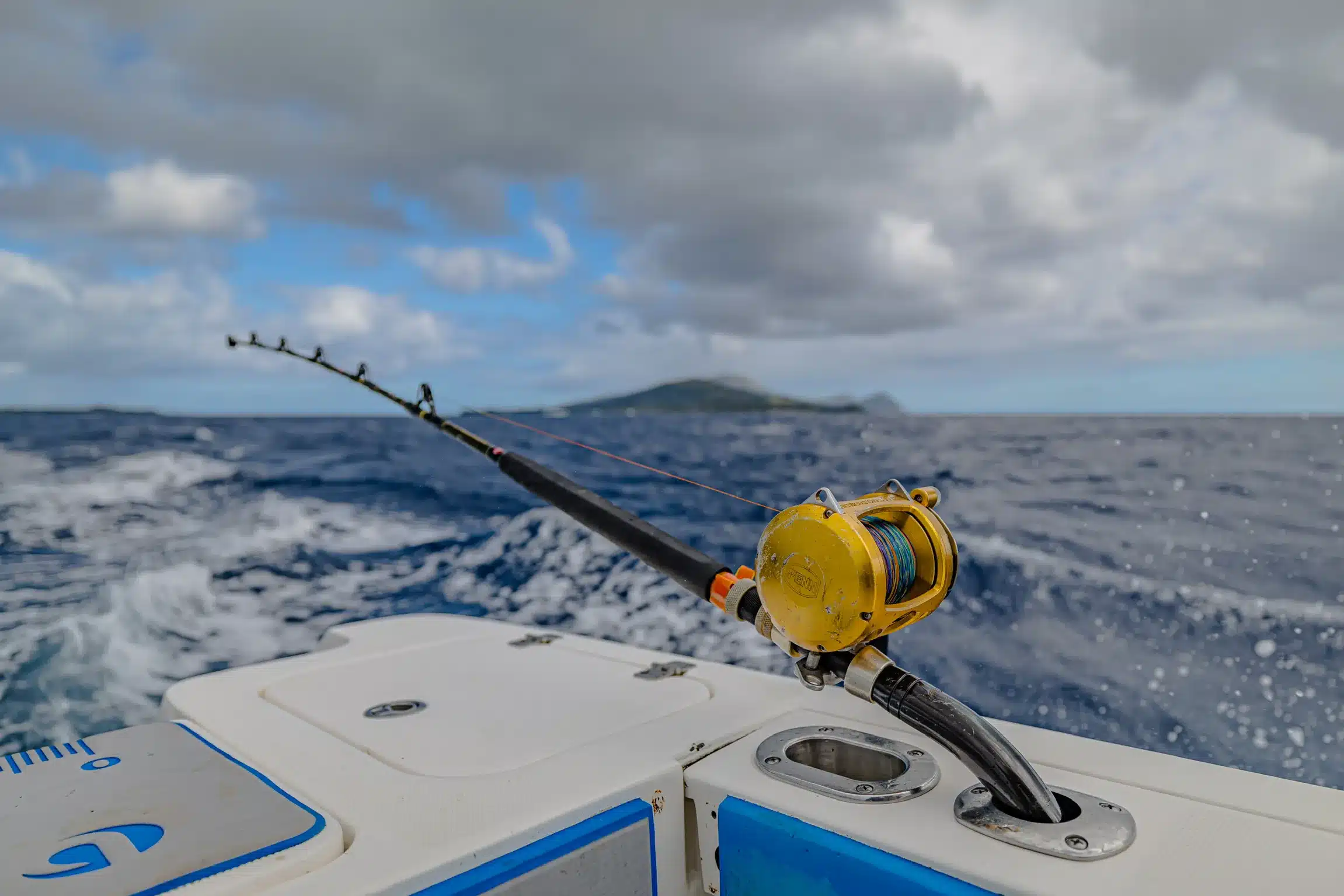Trolling rod with a gold game reel set in a rod holder on a boat in Vanuatu, with rough blue water and an island on the horizon.