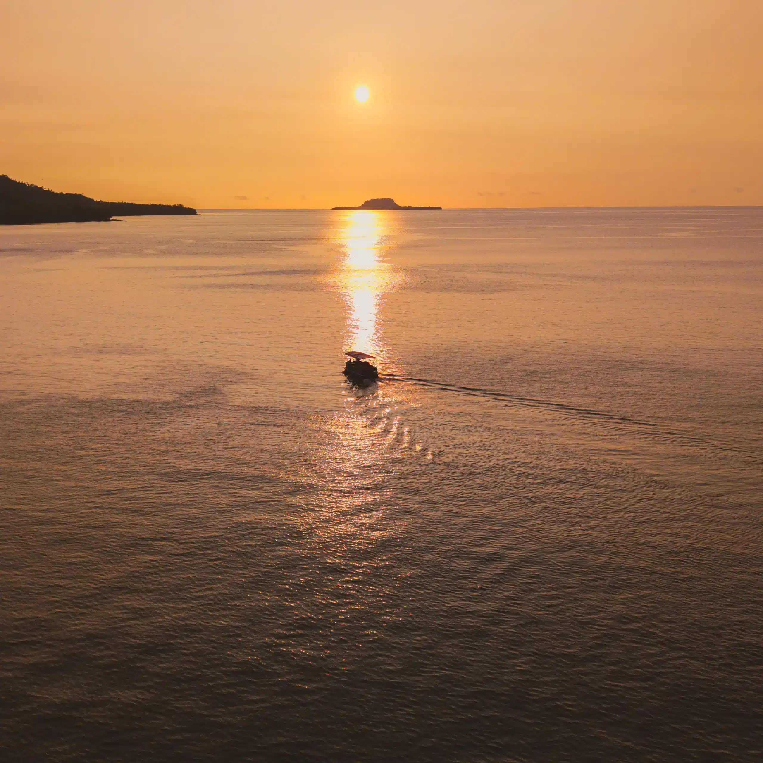 Boat cruising across calm water at sunset in Vanuatu, leaving a wake in golden light with islands on the horizon.