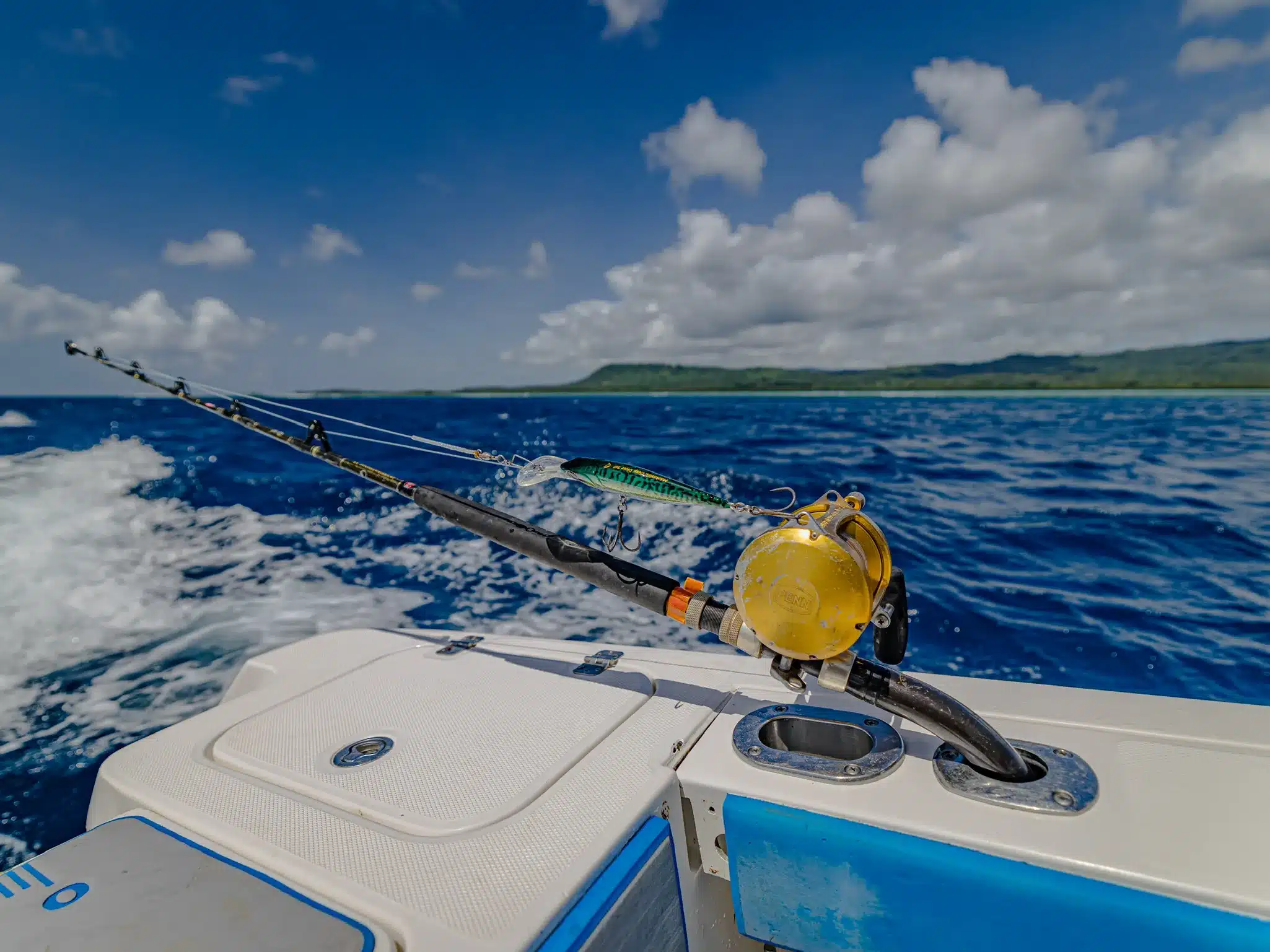 Trolling rod with a gold game reel and lure set out behind a boat on blue water in Vanuatu, with island coastline on the horizon.