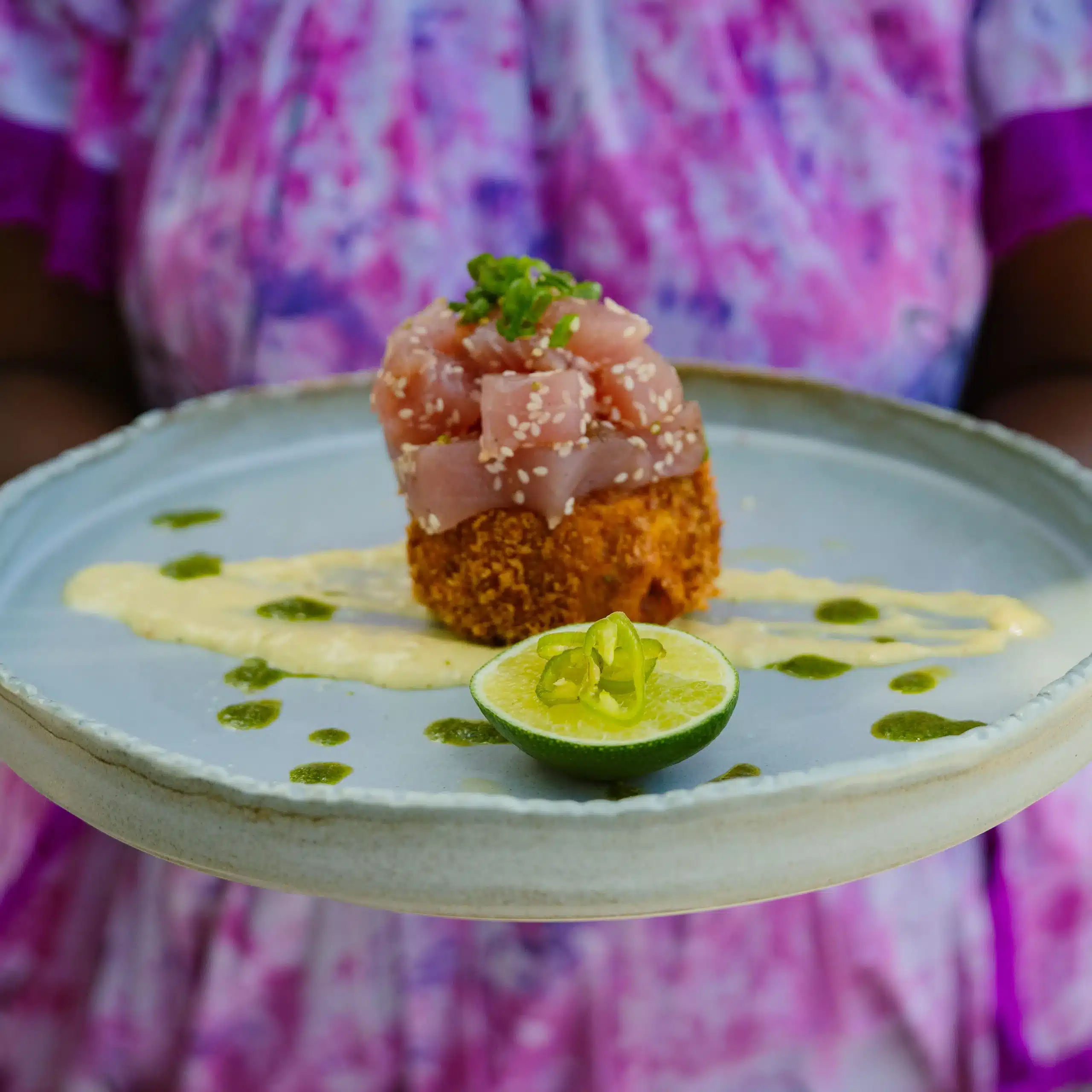 Plated tuna dish with diced raw tuna on a crispy base, garnished with sesame seeds and herbs, served with sauce and a halved lime.