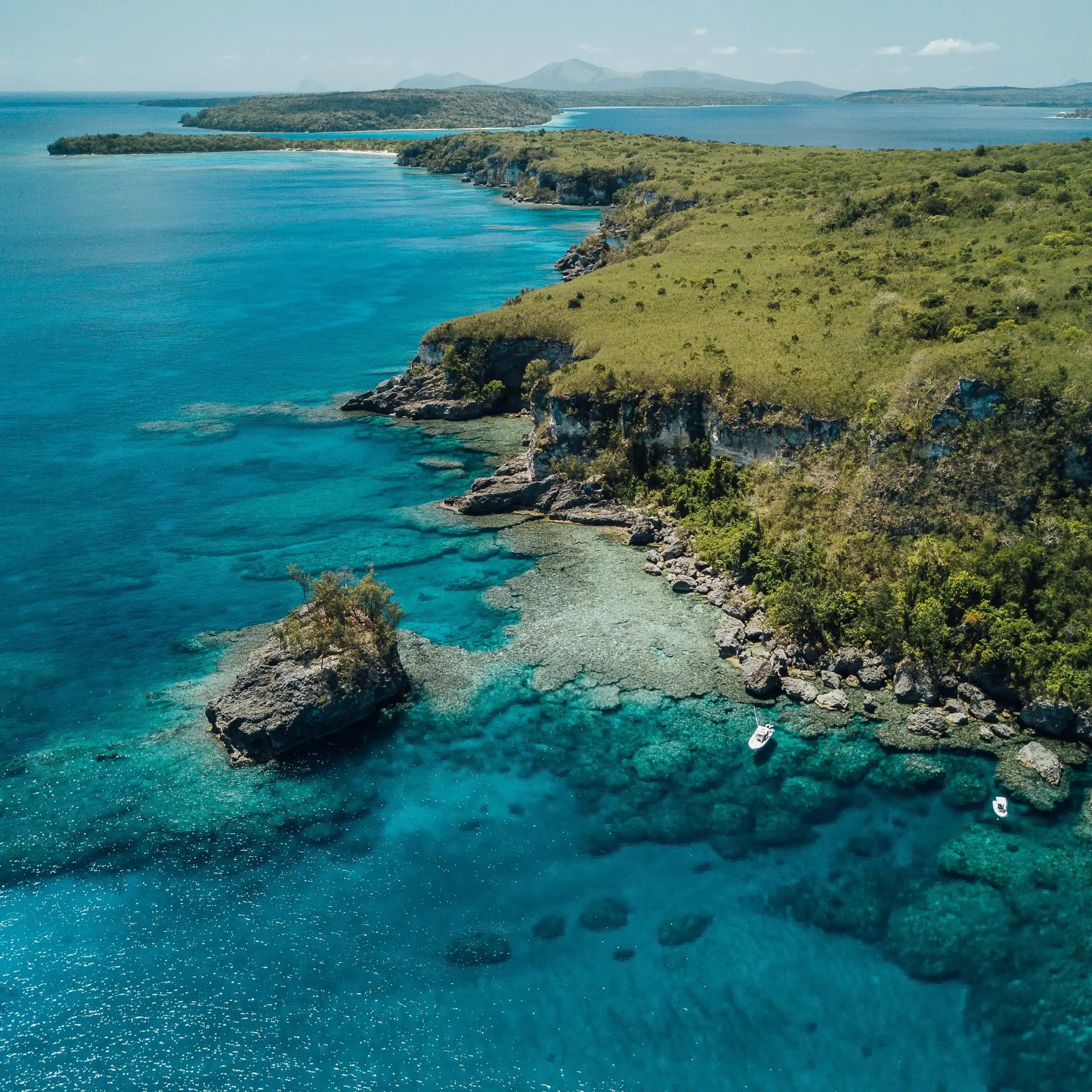 Aerial view of a rocky tropical coastline in Vanuatu with clear turquoise water over coral reef and two small boats anchored near shore.