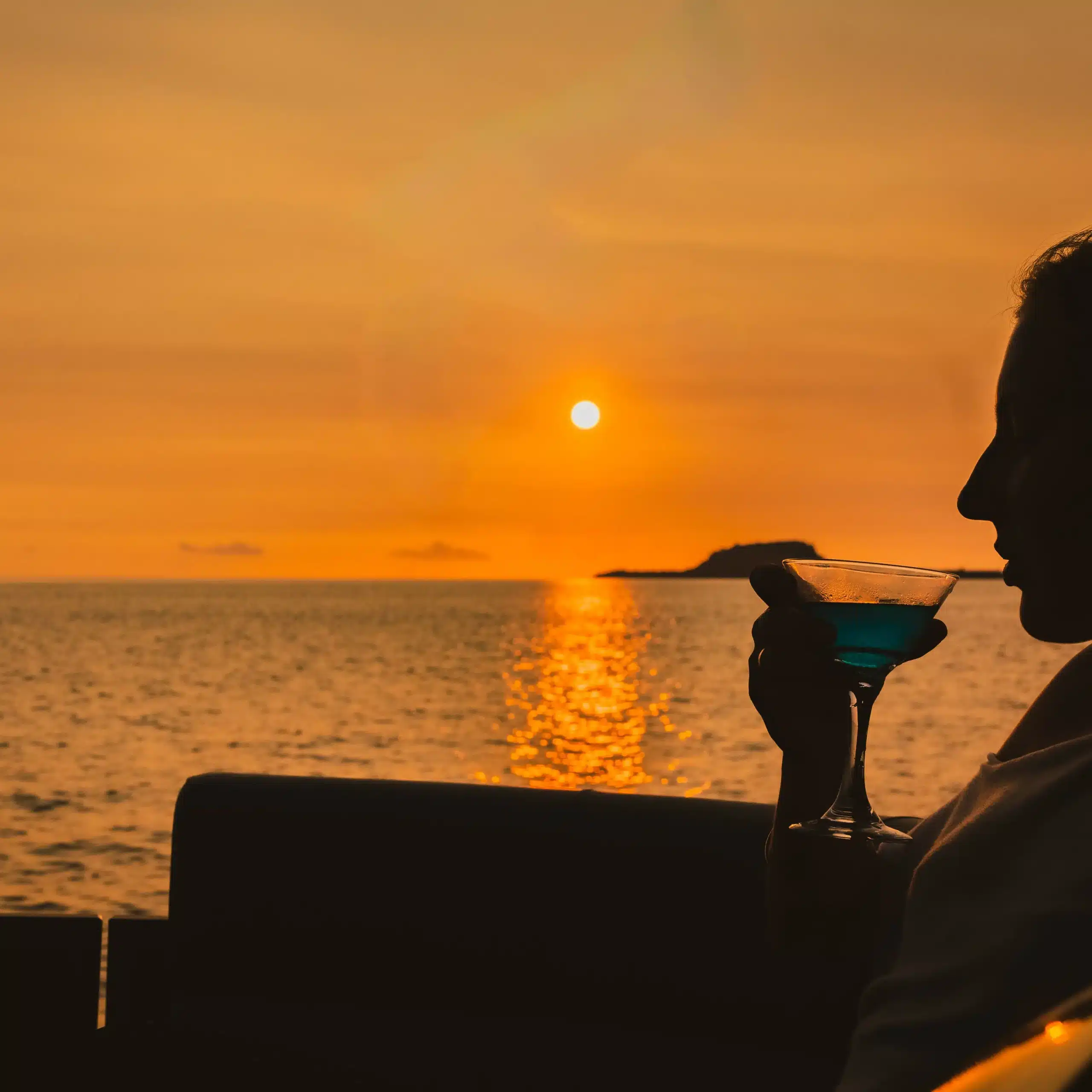Silhouette of a person holding a cocktail while watching a sunset over the ocean in Vanuatu, with golden light reflecting on calm water.
