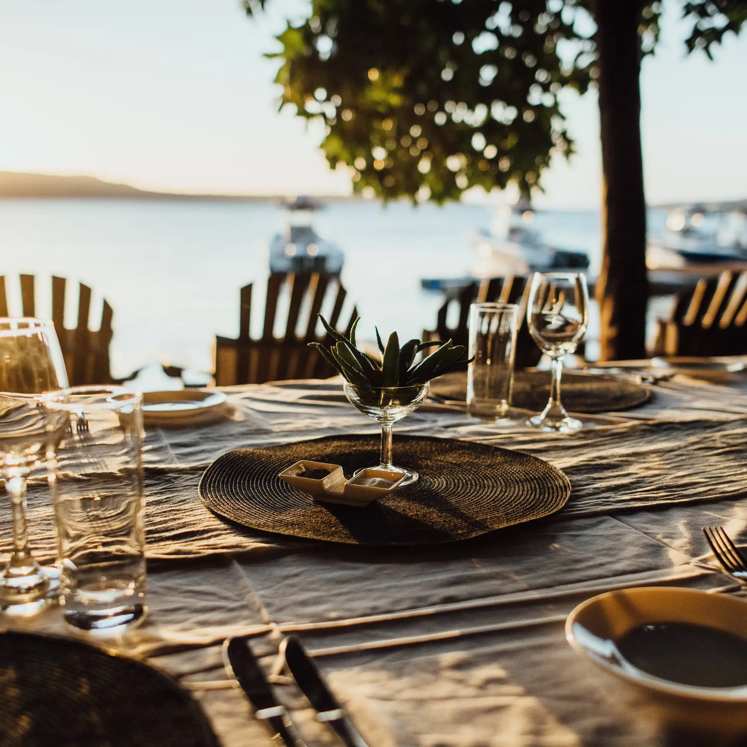 Outdoor dining table set for dinner at Trees and Fishes in Vanuatu, with wine glasses and place settings overlooking the waterfront at sunset.