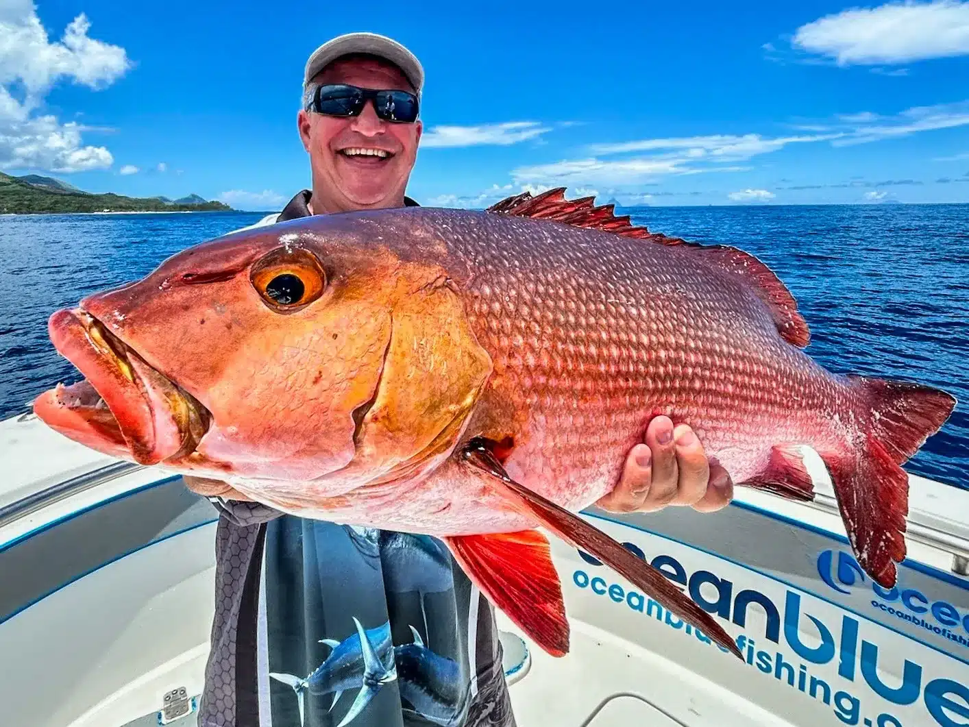 Smiling angler holding a large red emperor snapper on an Ocean Blue Fishing Adventures boat in Vanuatu, with blue ocean and coastline behind.